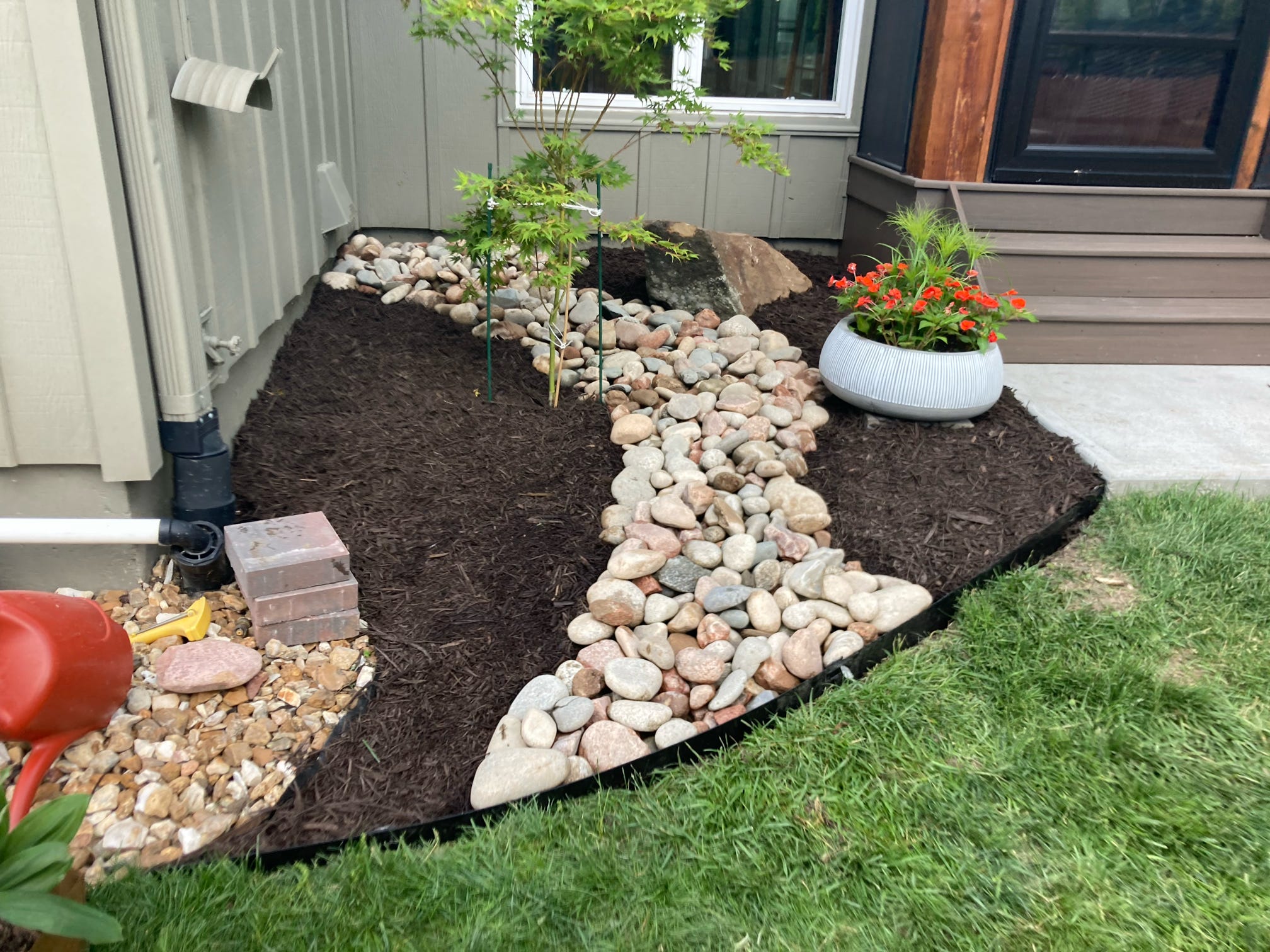 A small landscaped garden features stones, mulch, a potted plant with red flowers, and a tree near a house entryway.