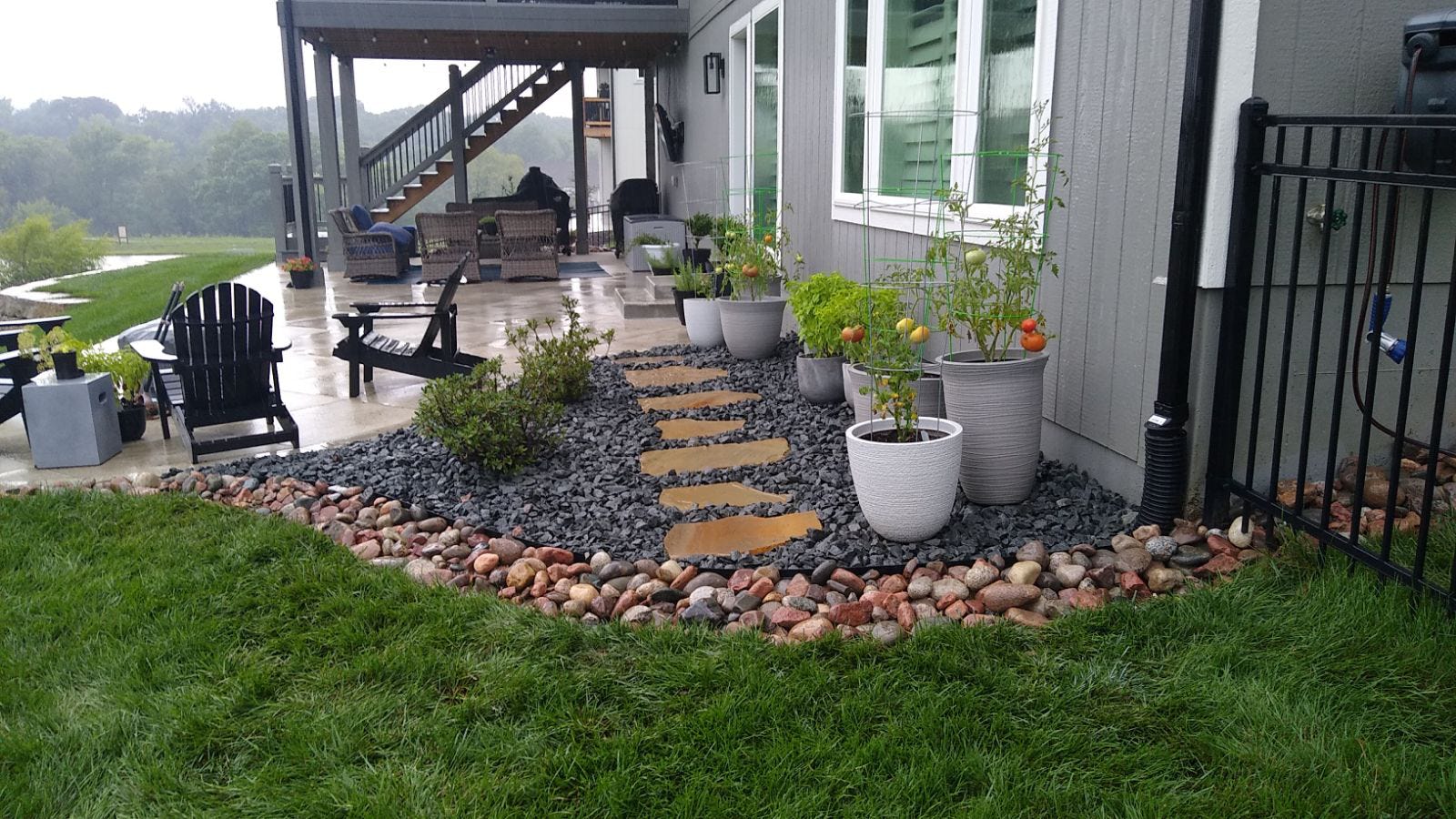 A cozy patio with Adirondack chairs, potted plants, and a stone path, overlooking a lush garden with trees in the background.