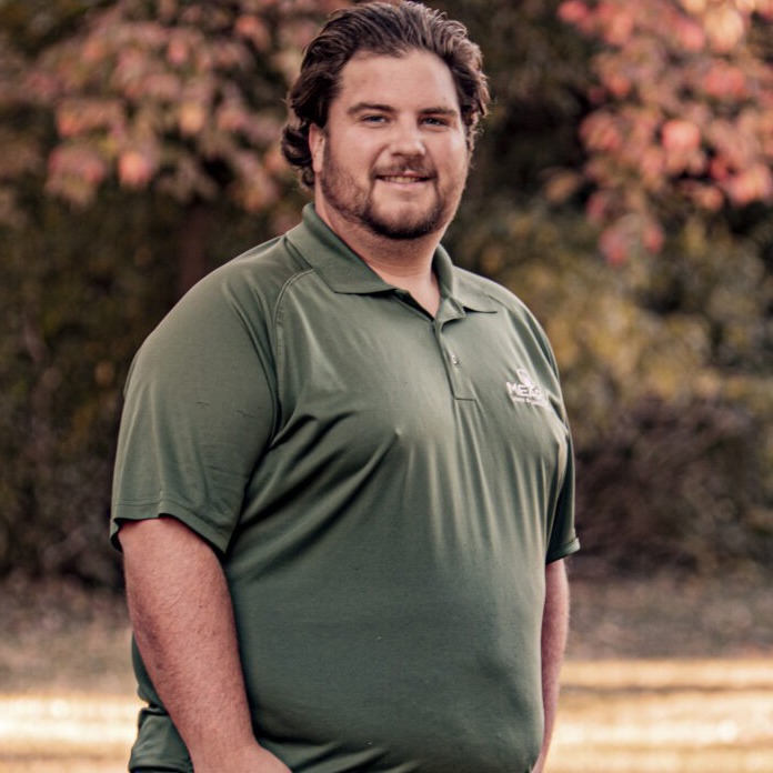 A person in a green polo shirt stands outdoors with blurred autumn foliage in the background, smiling at the camera.