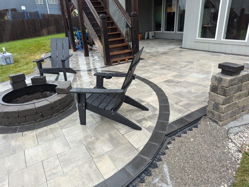 A patio with gray stone tiles features two Adirondack chairs by a circular brick fire pit. Stairs lead to the home's upper level.