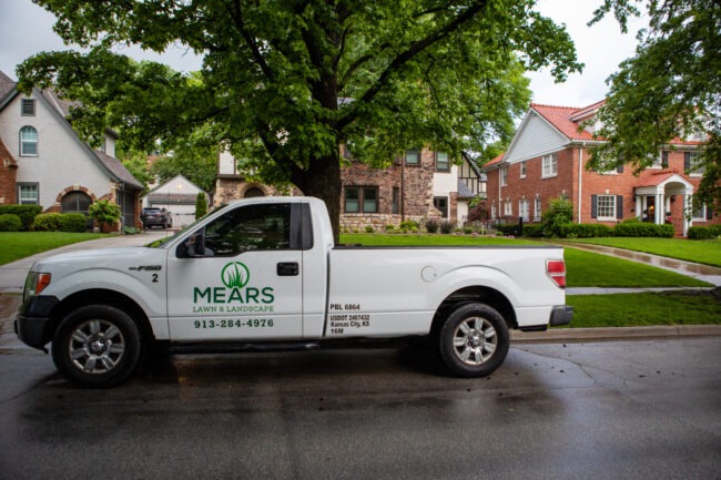 A landscaping truck is parked on a residential street with well-maintained lawns and traditional houses under a large, leafy tree.