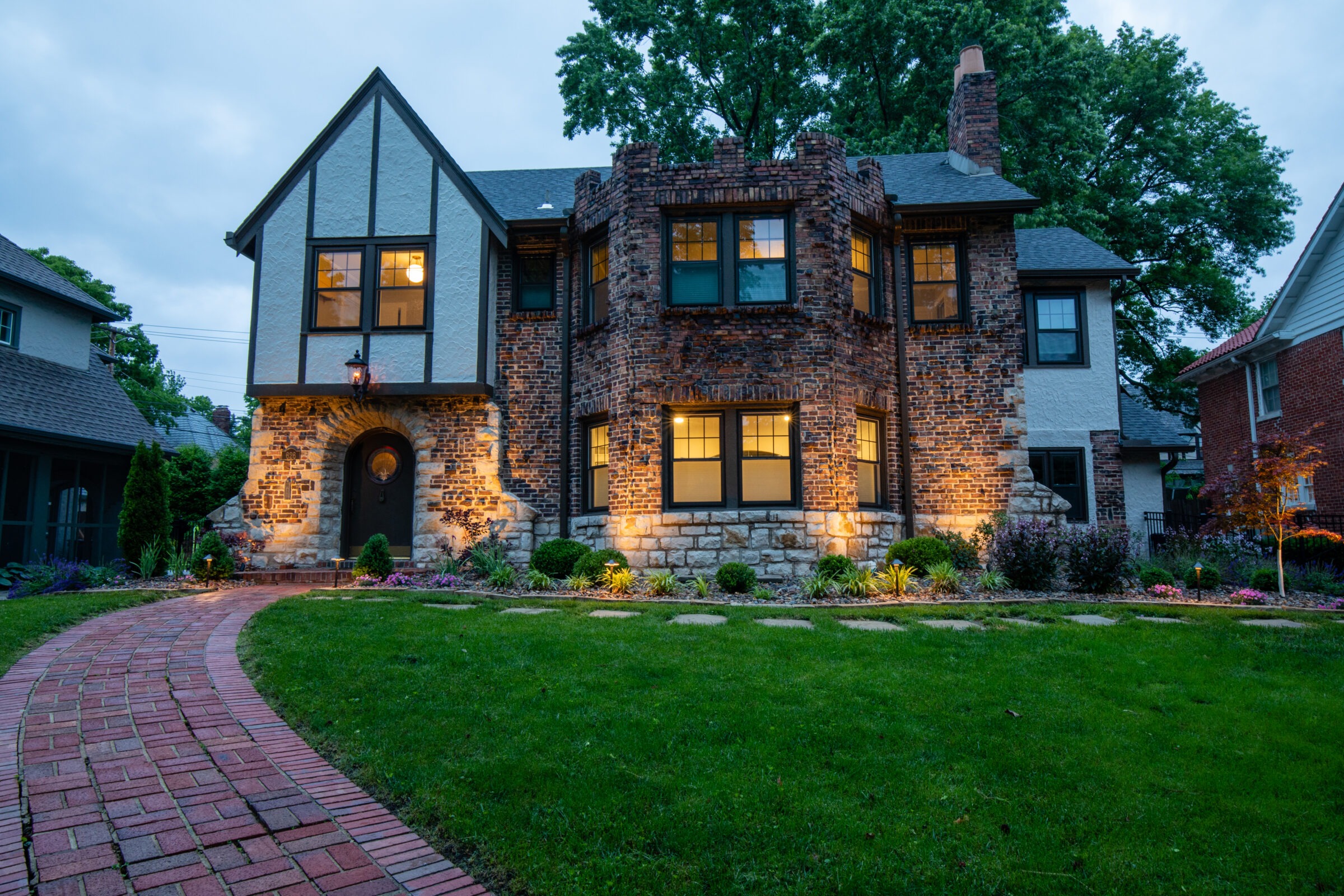 Tudor-style brick house with arched doorway, lit by exterior lights. Curved brick pathway, manicured lawn, and trees in the background. Evening setting.