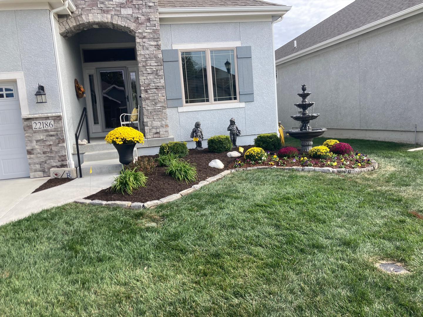 A neatly landscaped front yard with flowers, a fountain, and a stone entrance. House has light blue siding and brick accents.