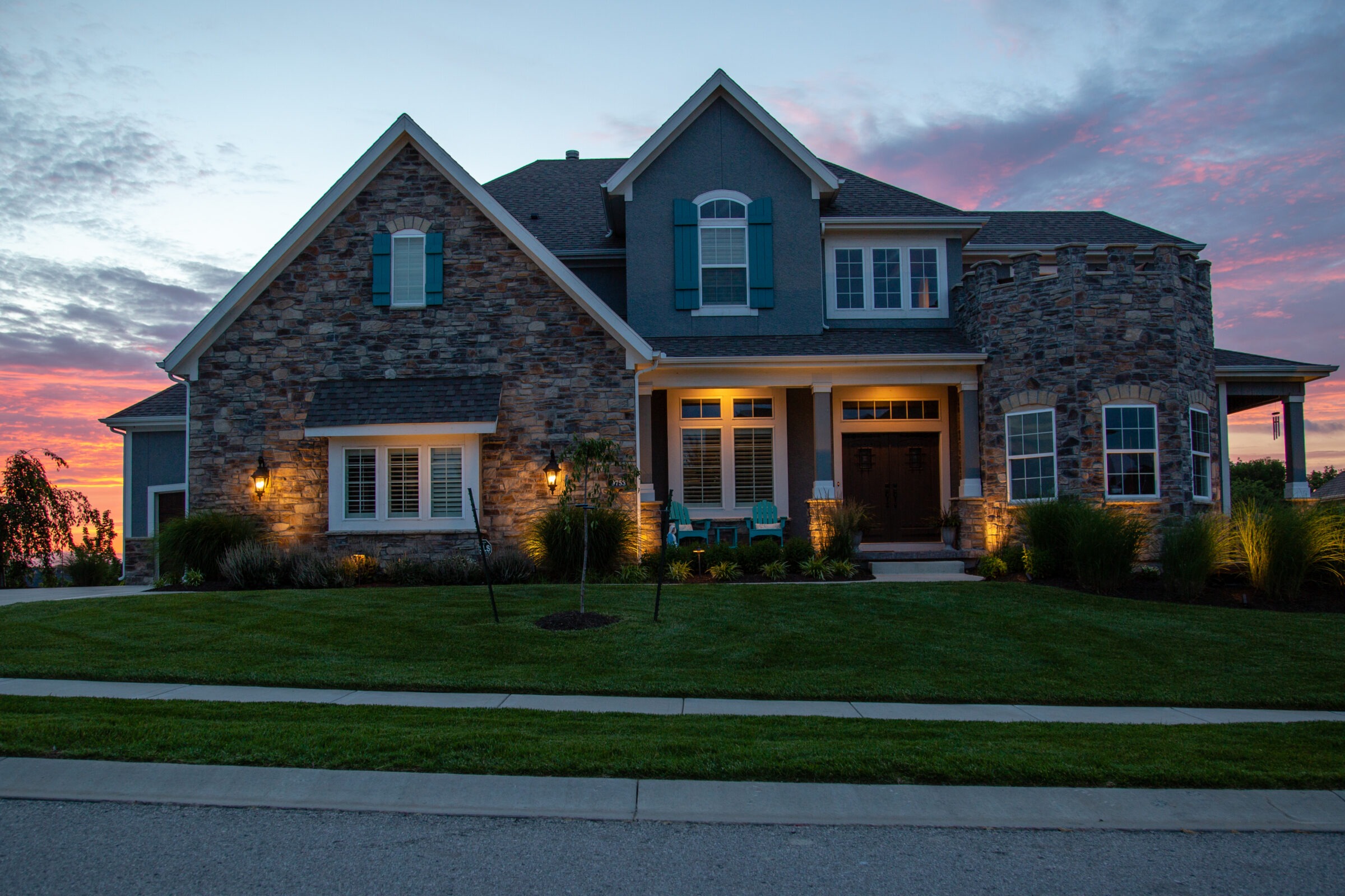 Elegant stone house at sunset, illuminated exterior lighting, blue shutters, green lawn, paved driveway, under a vibrant, colorful sky.