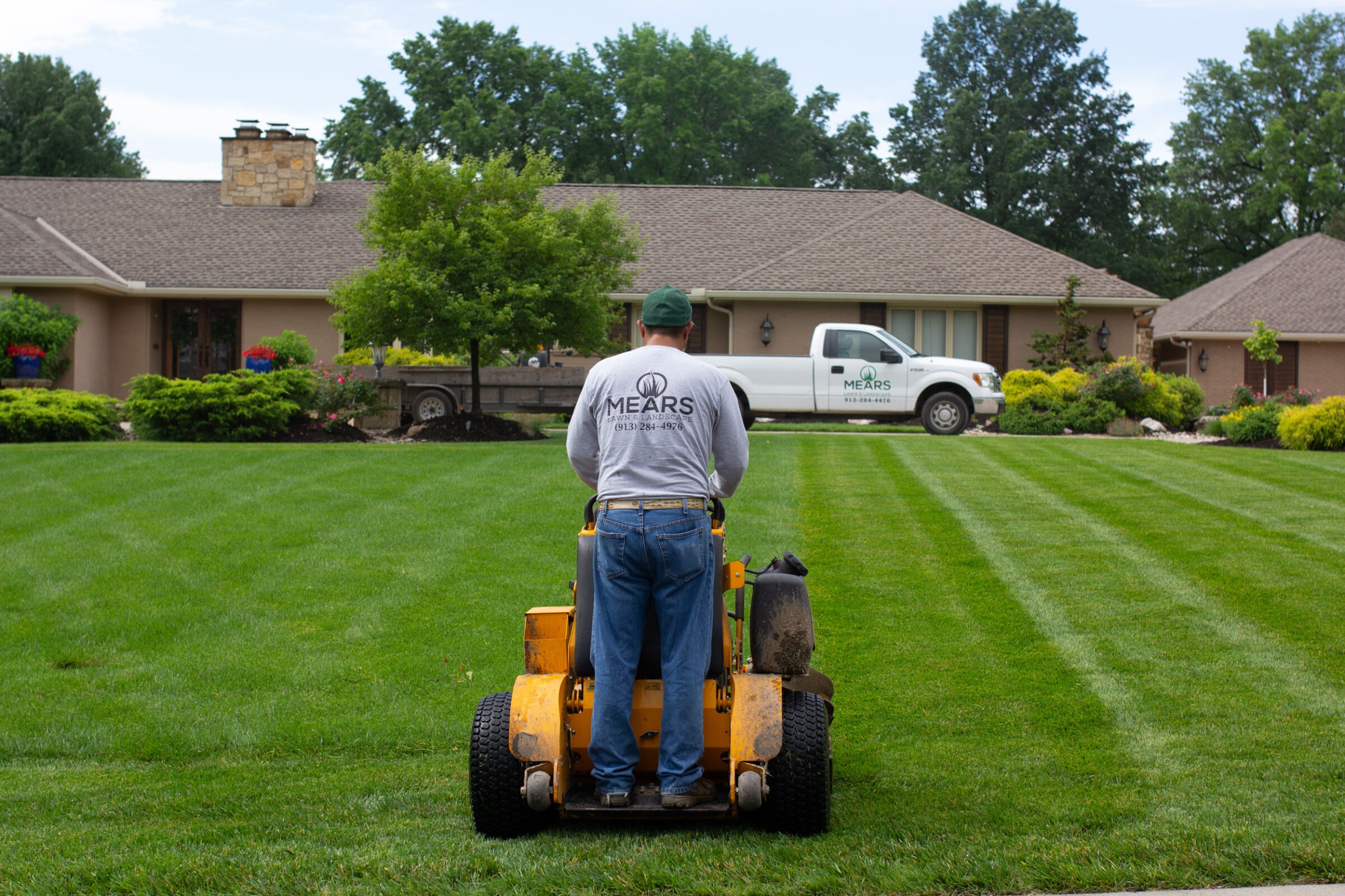 A person operates a lawn mower on a neatly manicured lawn, with a landscaping truck and house in the background.