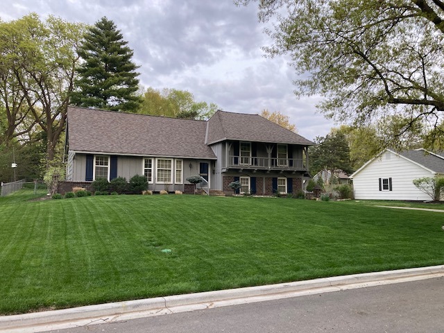 A suburban two-story house with a well-maintained lawn, surrounded by trees and neighboring homes under an overcast sky. No people are visible.