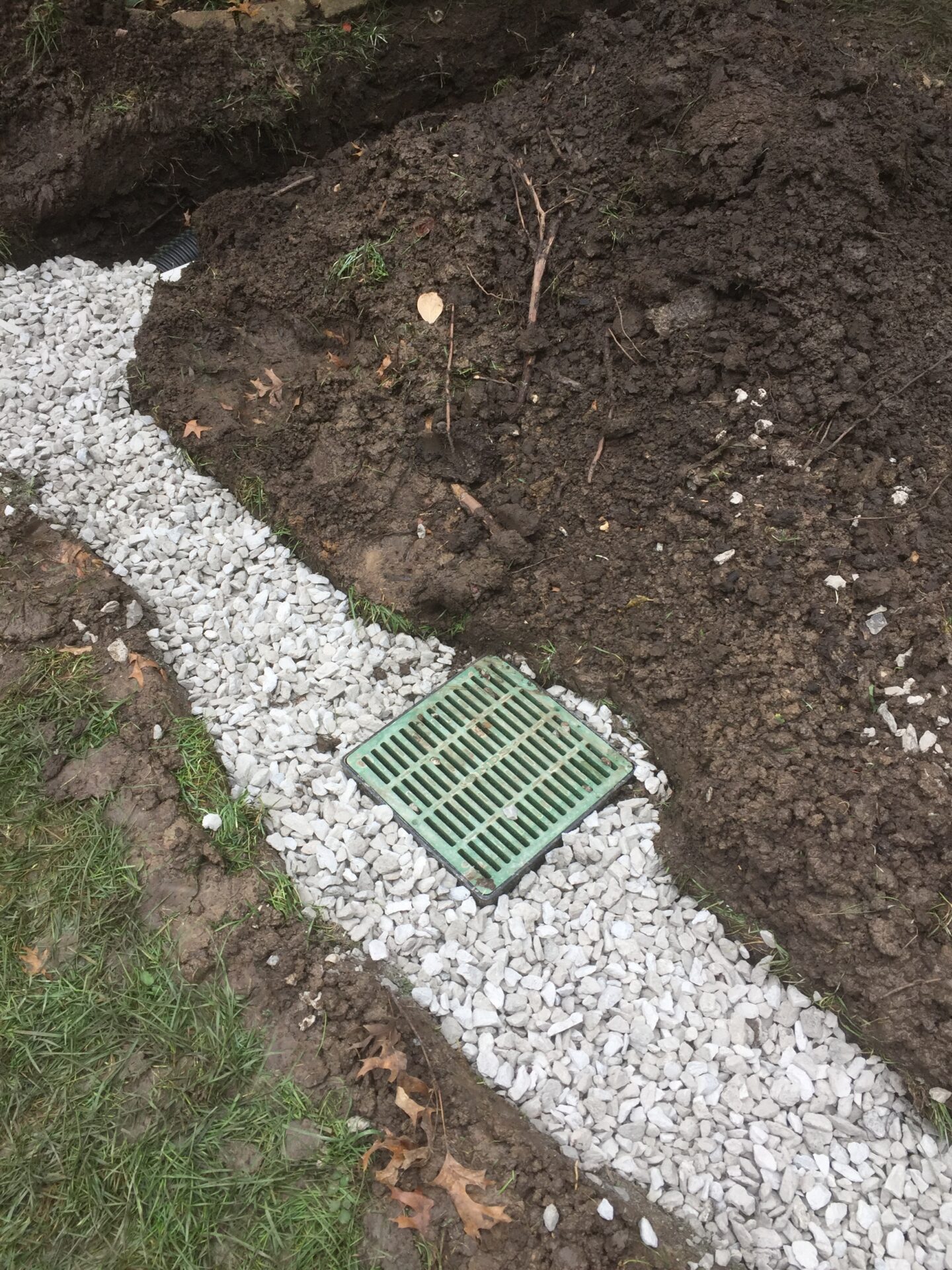 A drainage system is being installed with rocks and a green grate partially buried in soil and surrounded by grass.