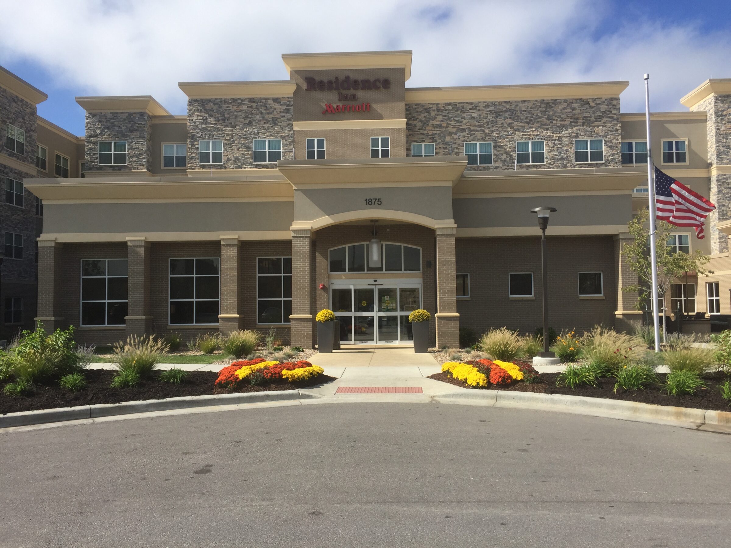 Hotel with stone facade and landscaped entrance. American flag on pole, clear sky above. Central front doors below Residence Inn signage.