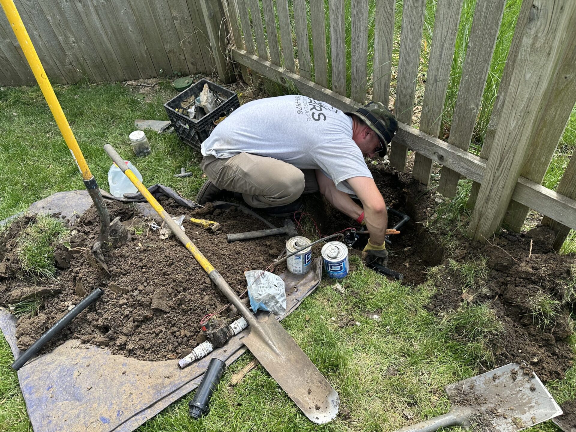 A person is repairing a pipe near a wooden fence, surrounded by tools, dirt, and grass in a backyard setting.