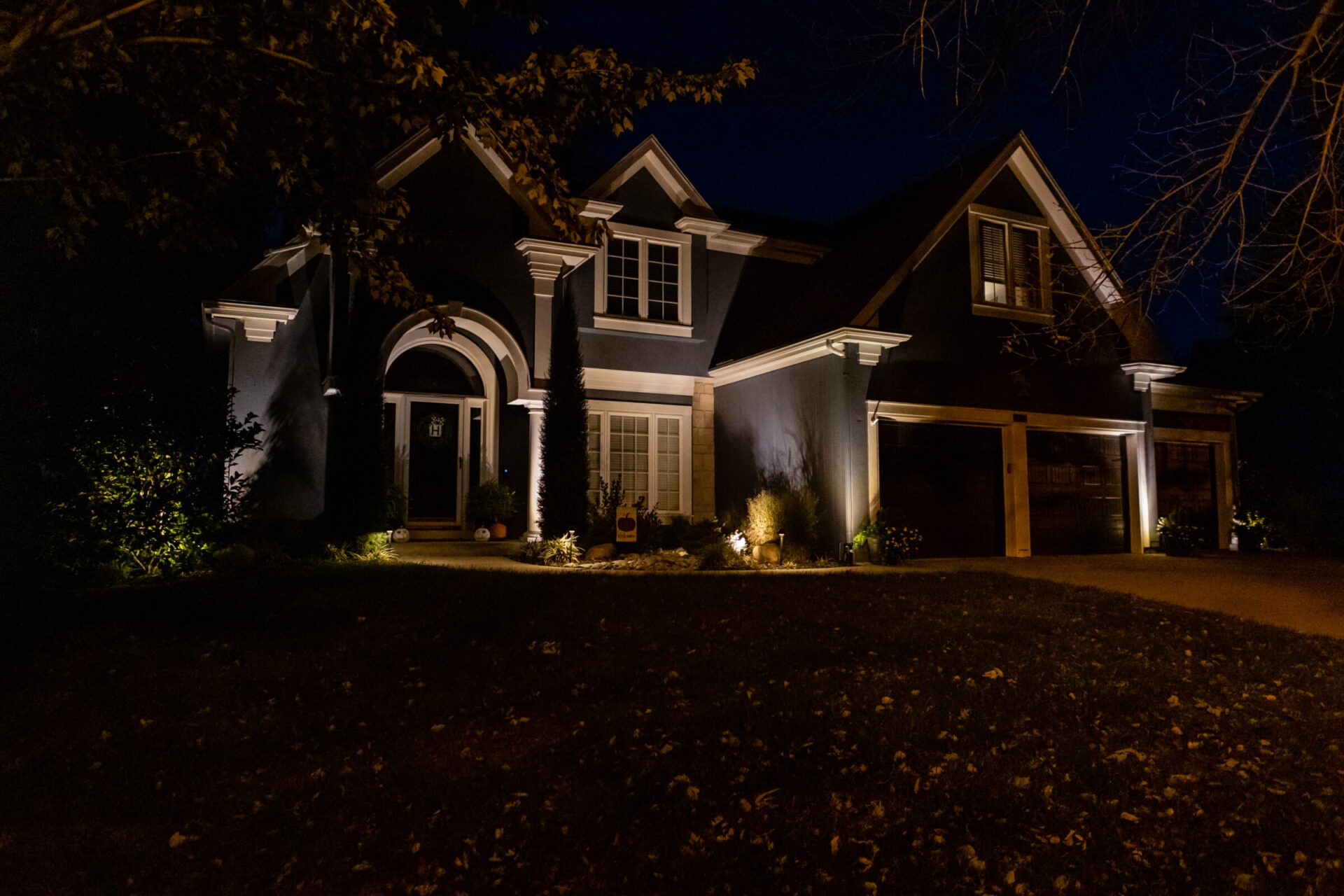Illuminated house at night with landscaping and a tree in the foreground, featuring a prominent arched doorway and garage doors.