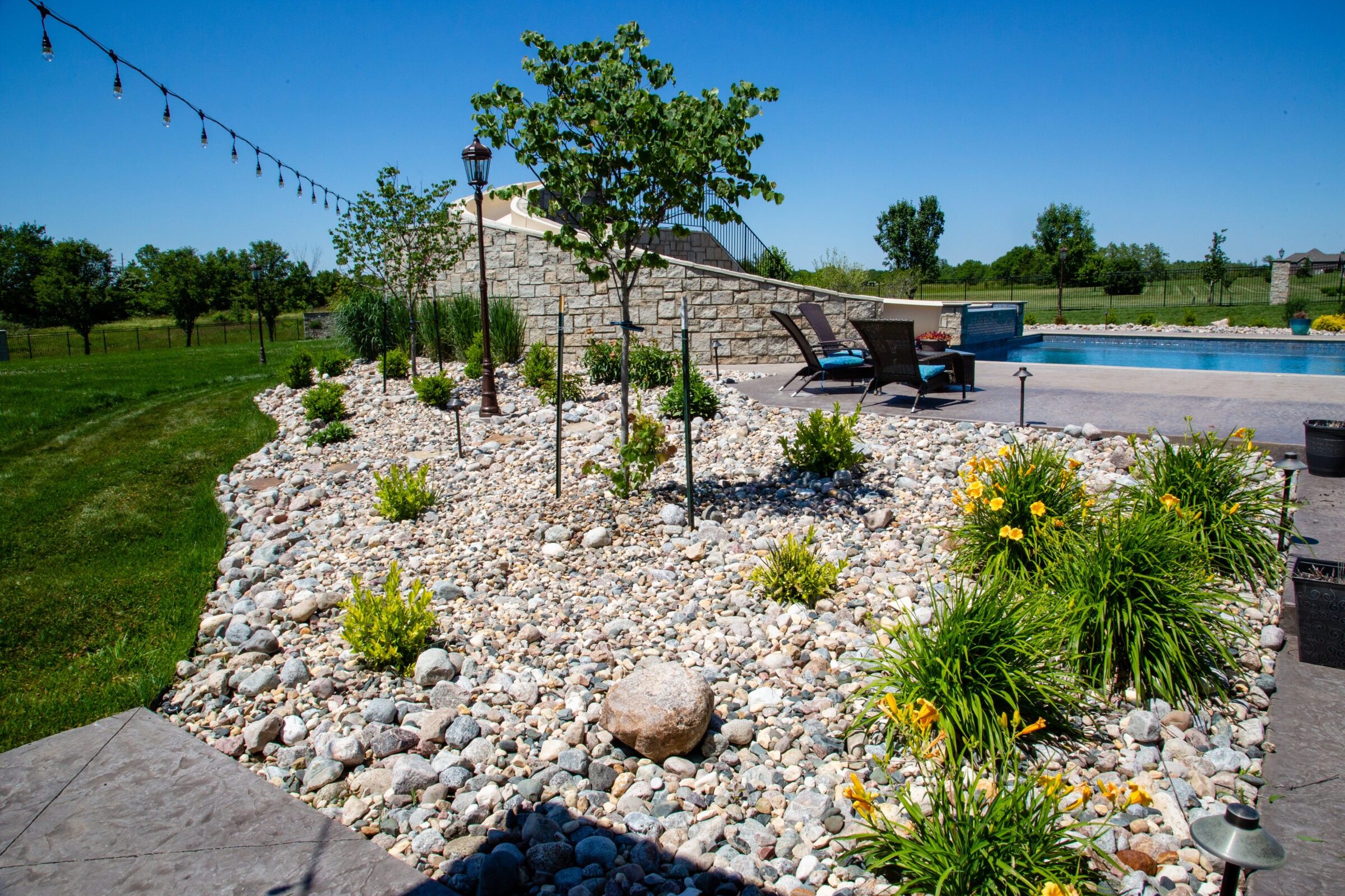 A landscaped backyard with a pool, stone path, string lights, and lounge chairs, surrounded by grass and trees under a clear blue sky.