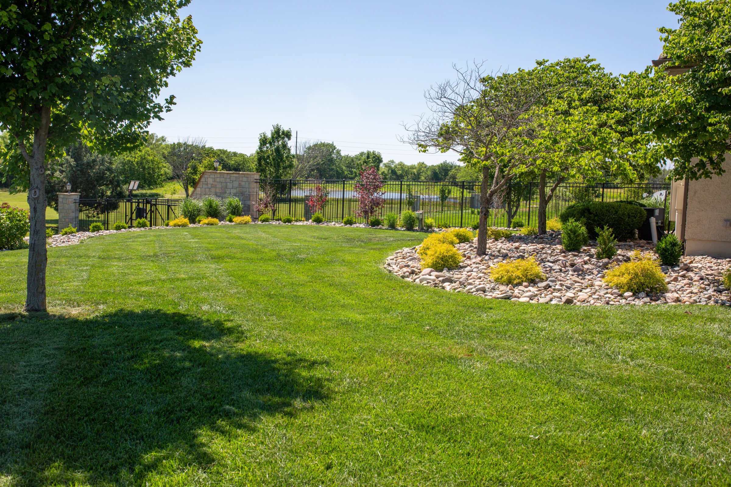 A well-maintained backyard with green grass, decorative stones, trees, and shrubs, bordered by a black metal fence and stone wall.