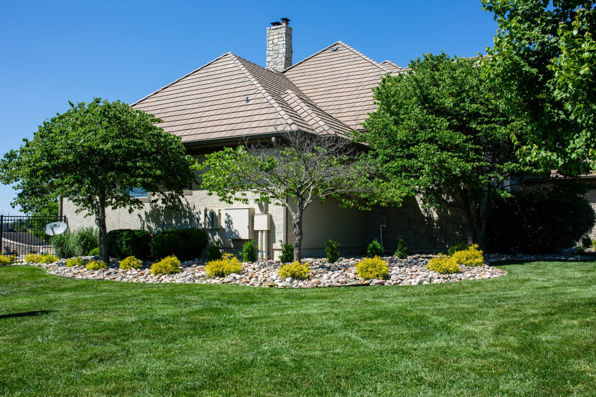 Suburban house with a neatly landscaped yard, surrounded by green grass, trees, and a rock garden under a clear blue sky.