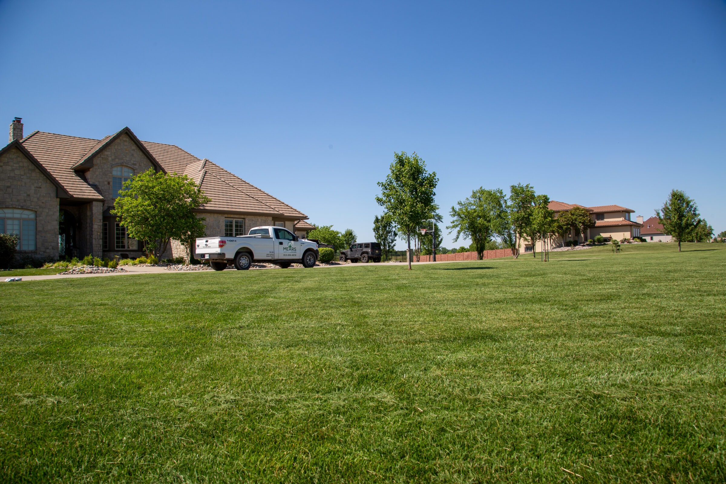 A spacious green lawn fronts a large suburban house with two vehicles parked outside, under a clear blue sky.