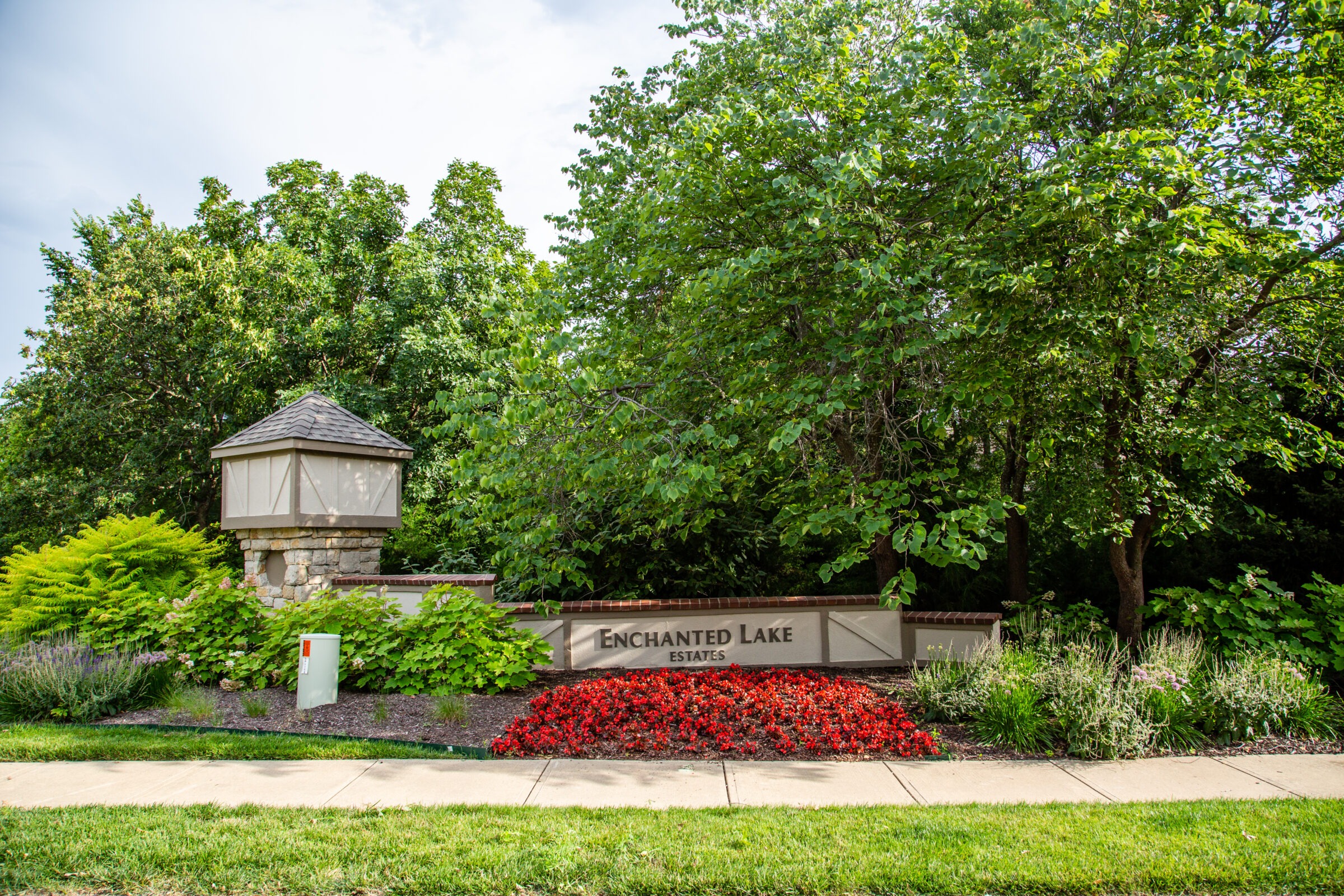 A landscaped entrance sign for Enchanted Lake Estates, surrounded by trees and red flowers, under a partly cloudy sky.