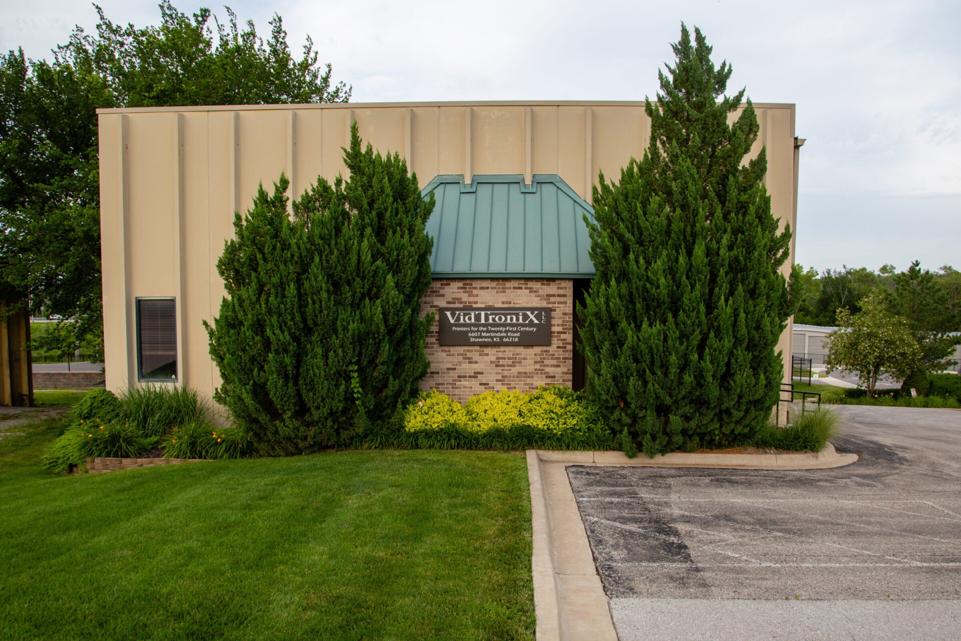 A beige building with green roof, surrounded by trees and grass, displays the sign 