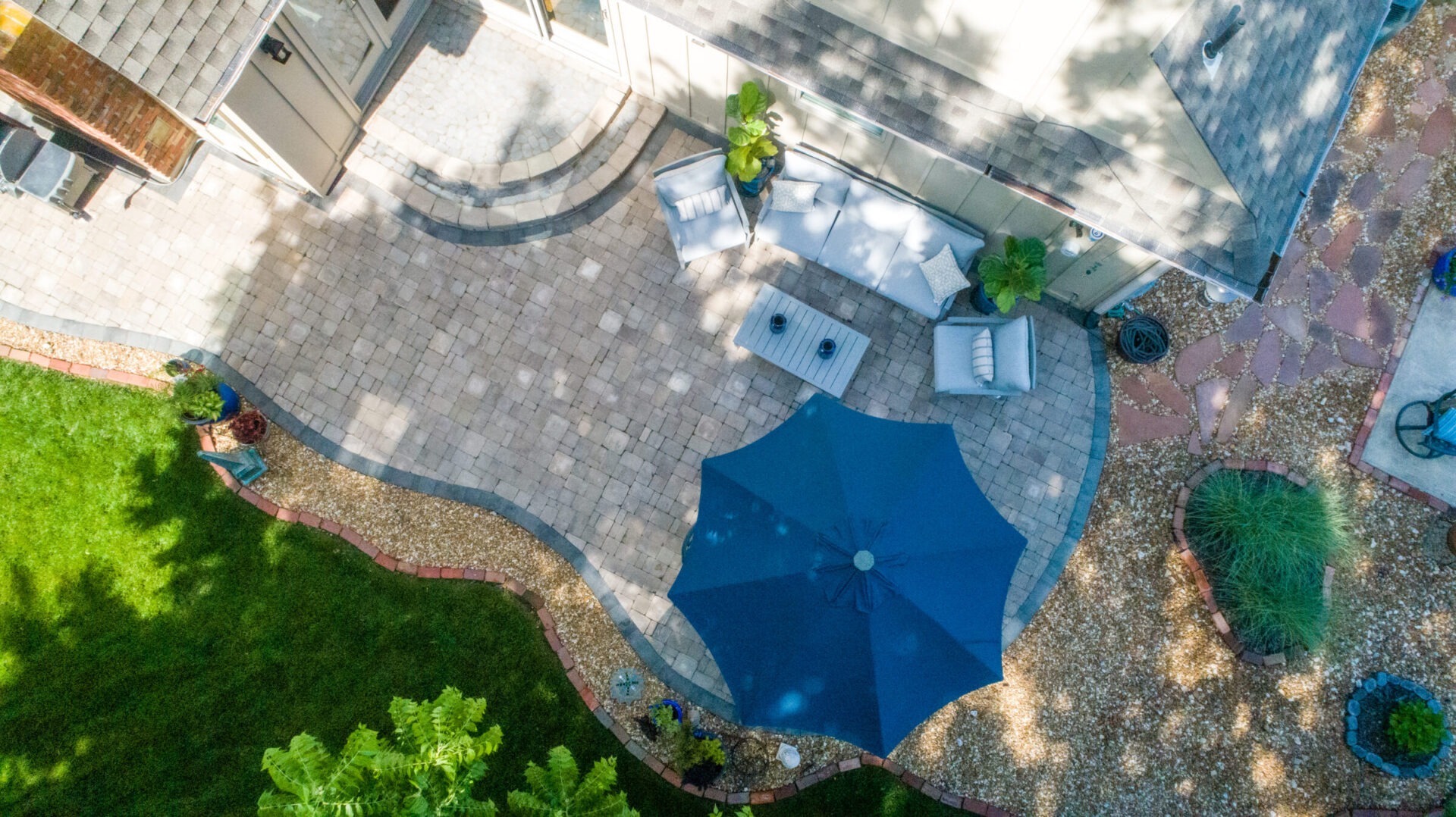 Aerial view of a patio with blue umbrella, seating area, and greenery, adjacent to a house. Shadows create a patterned effect.