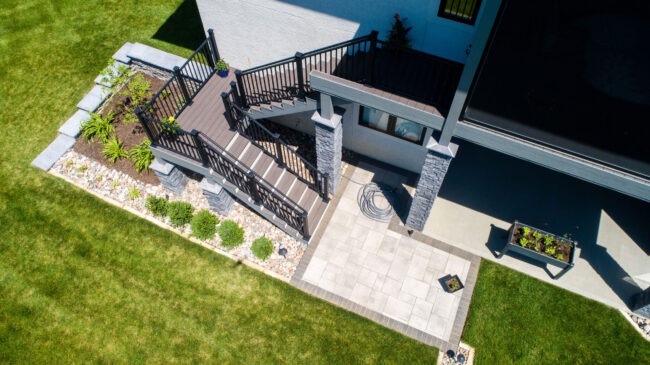 Aerial view of a modern backyard with a staircase, stone patio, neatly trimmed lawn, and landscaped garden with decorative plants and edging.