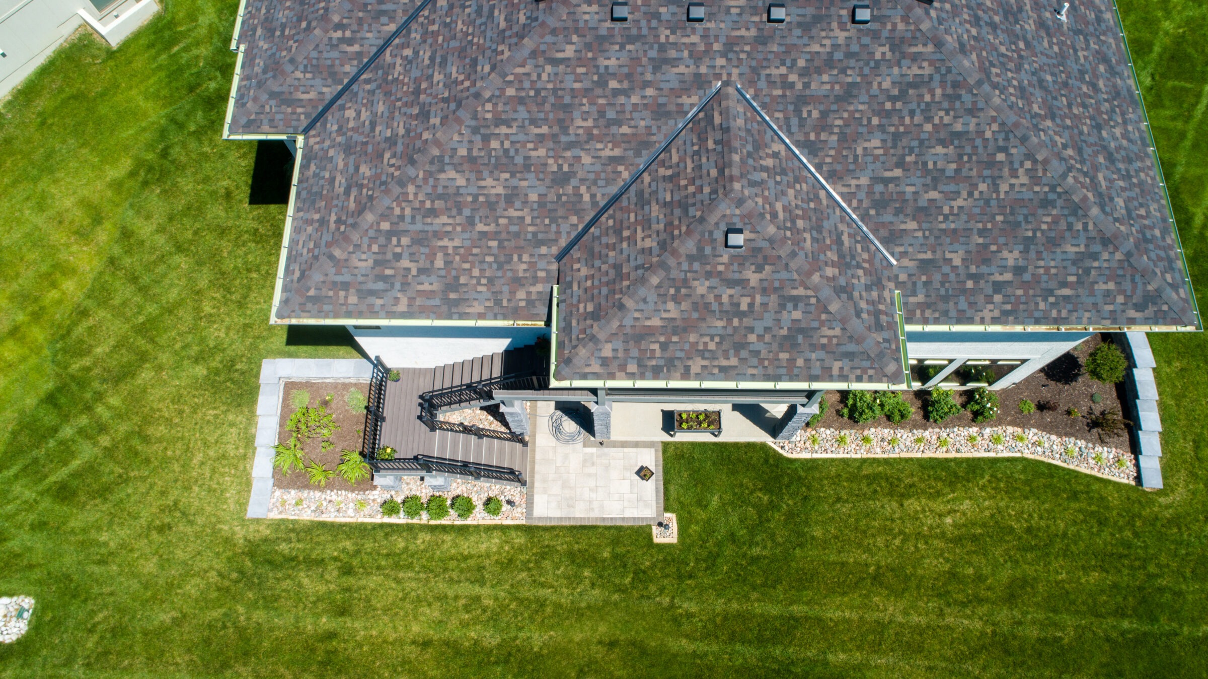 Aerial view of a house with a patterned roof, surrounded by neatly trimmed grass and landscaped gardens, featuring a patio and staircase.