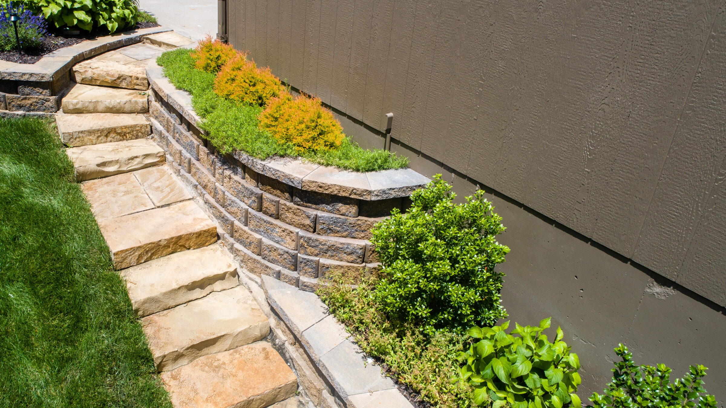 Stone steps lead up a landscaped garden path alongside a wooden wall, with greenery and plantings on the right side.