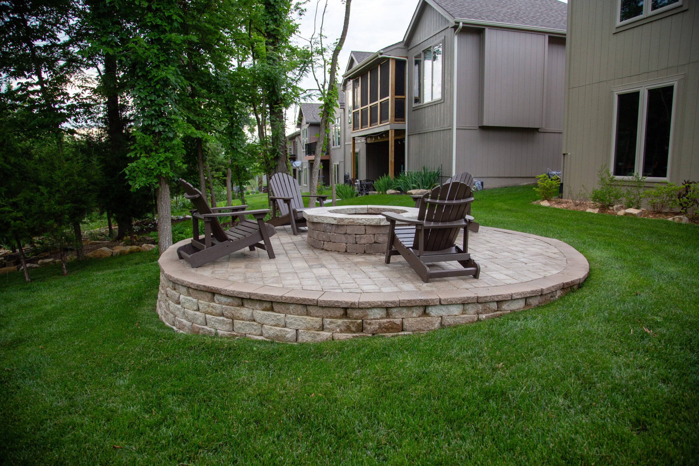 Circular patio with Adirondack chairs encircling a fire pit, surrounded by green grass and trees, adjacent to a modern suburban house.