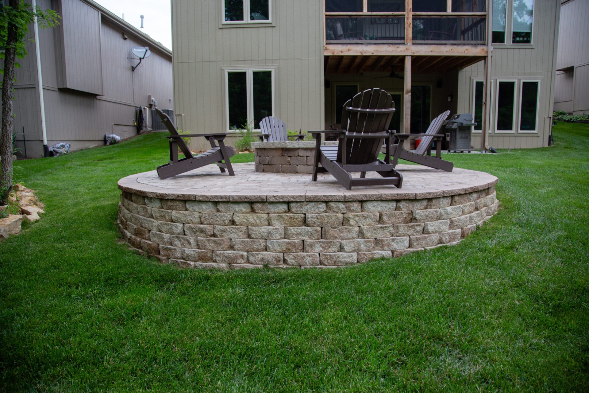 A backyard scene with a stone patio, adirondack chairs, and fire pit, surrounded by grass, adjacent to a beige two-story house.