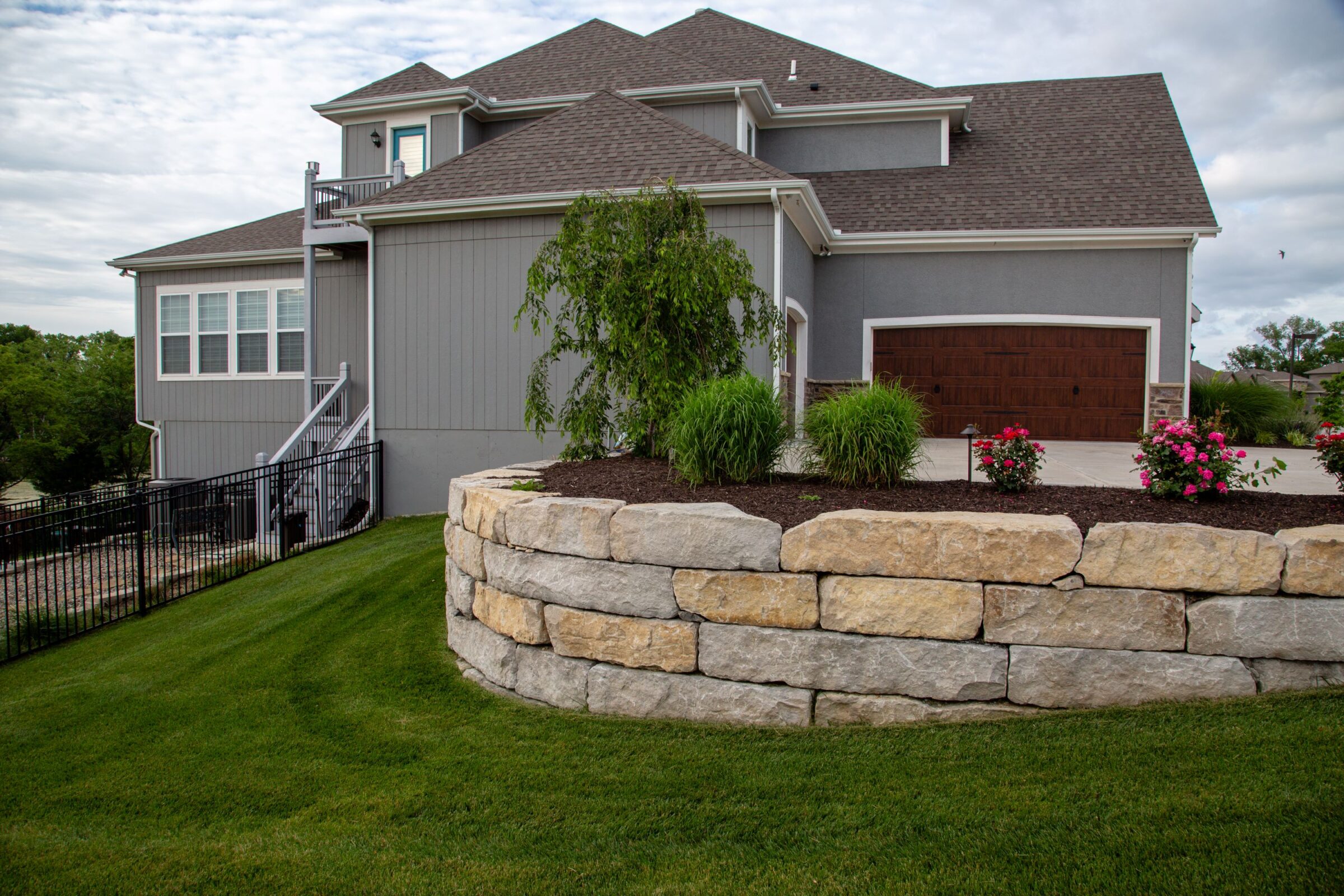 Gray house with brown garage, surrounded by landscaped yard. Stone retaining wall and colorful flowers in foreground. Cloudy sky above.