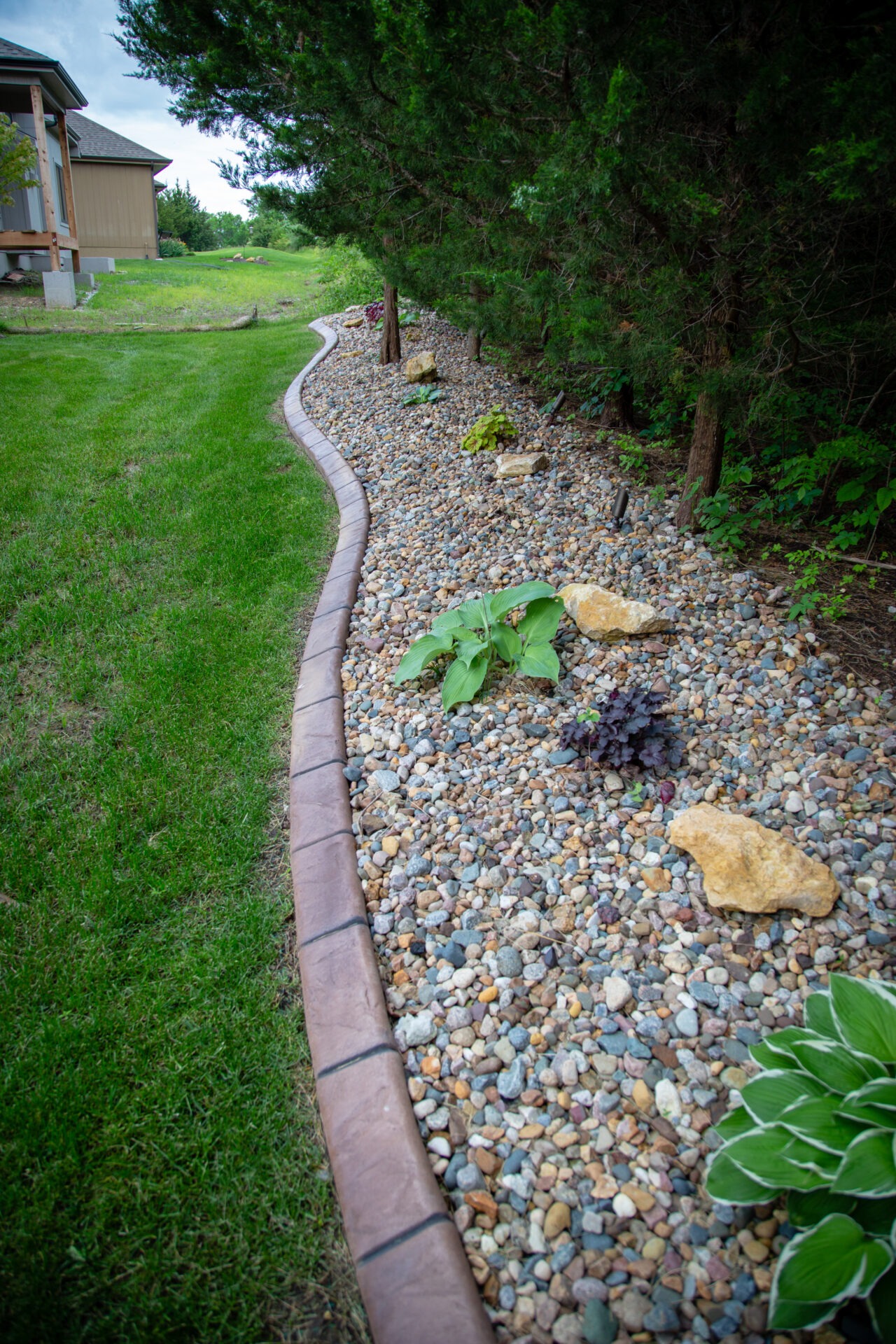 A landscaped garden with curved stone edging, featuring gravel, various plants, and green grass, bordered by lush trees and a building.