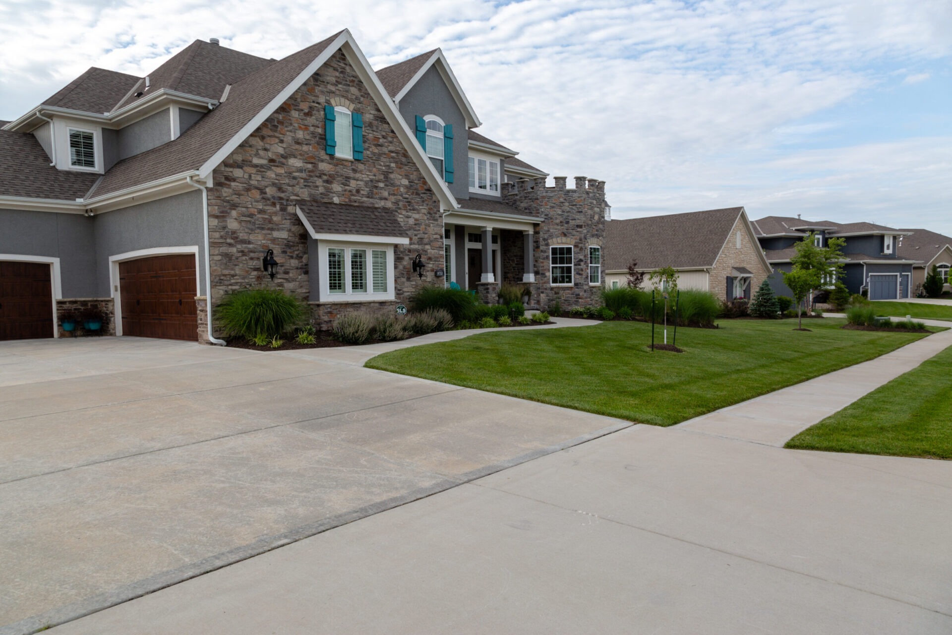 A spacious suburban house with stone facade and teal shutters, surrounded by neatly landscaped lawns and a wide driveway under a partly cloudy sky.