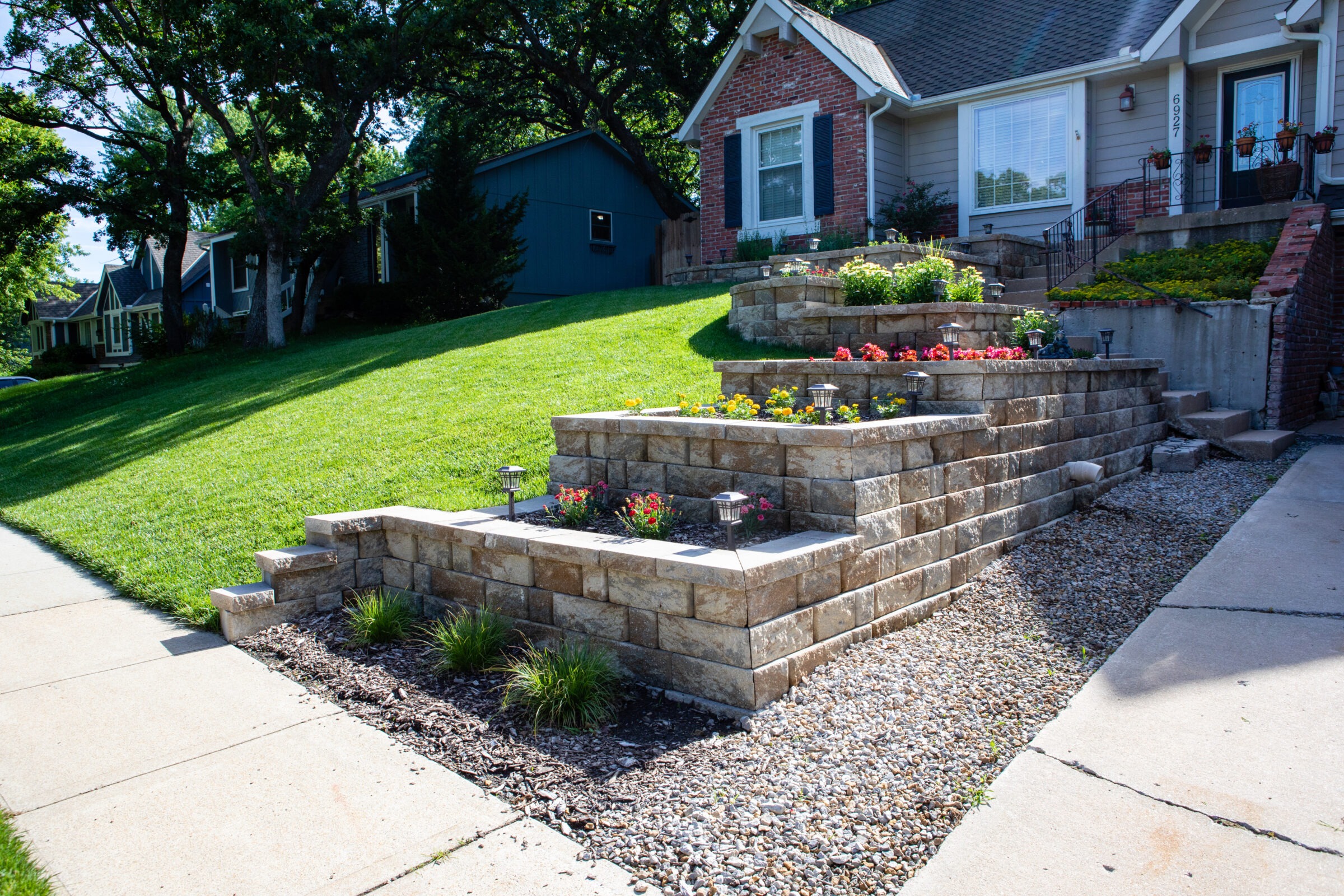 Neatly landscaped front yard with tiered stone walls and colorful flowers. Adjacent houses surrounded by lush green lawn and sidewalk.