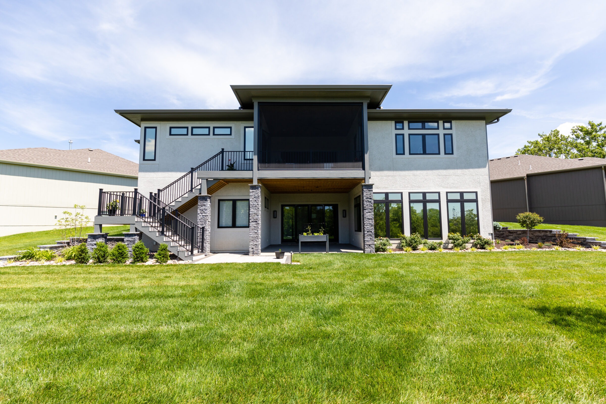 Modern two-story house with large windows, surrounded by a neatly manicured lawn. No people visible, bright sunny day, suburban neighborhood.