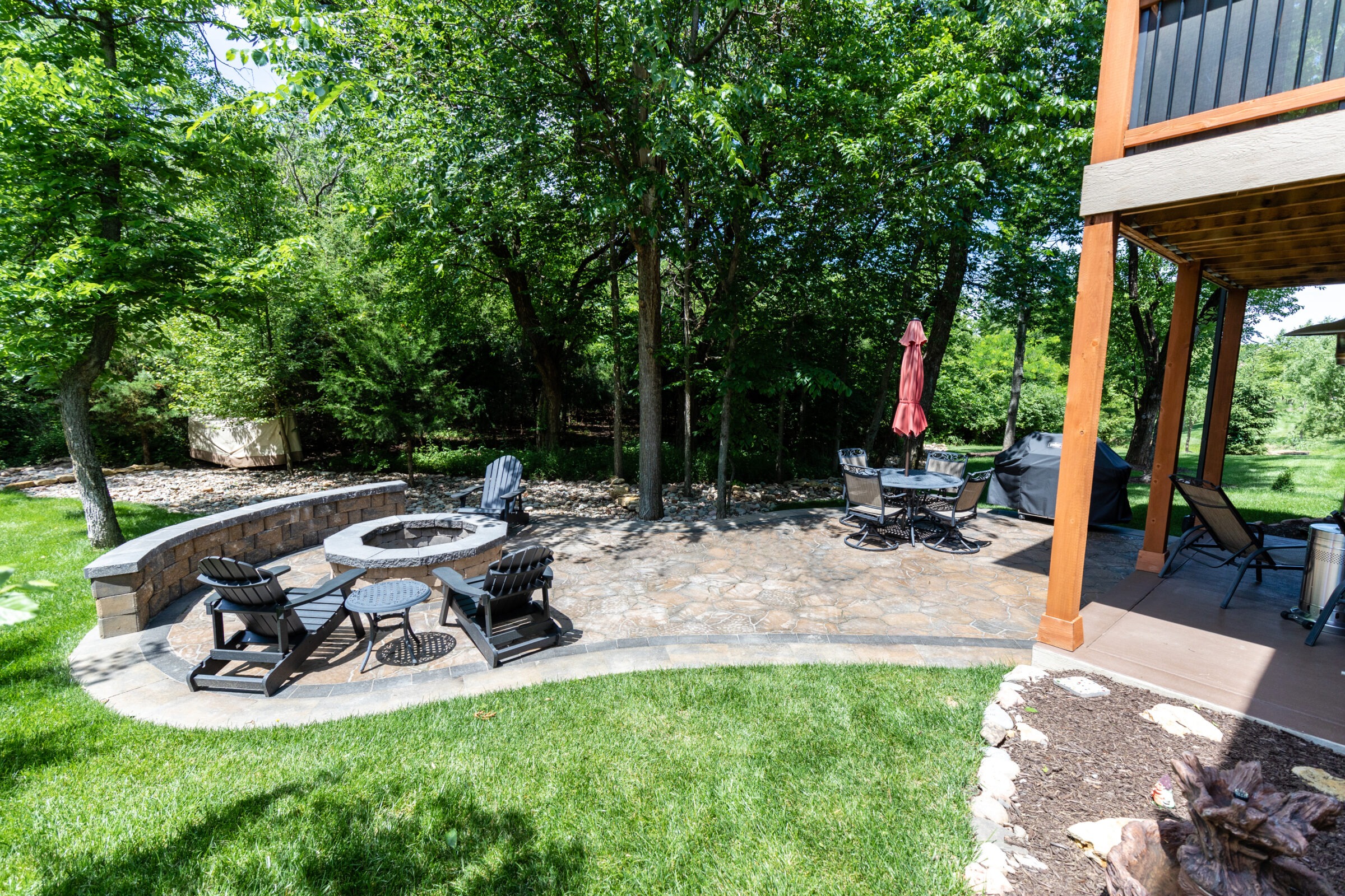 A sunny patio features Adirondack chairs around a fire pit, dining table with umbrella, and a grill, surrounded by lush green trees.