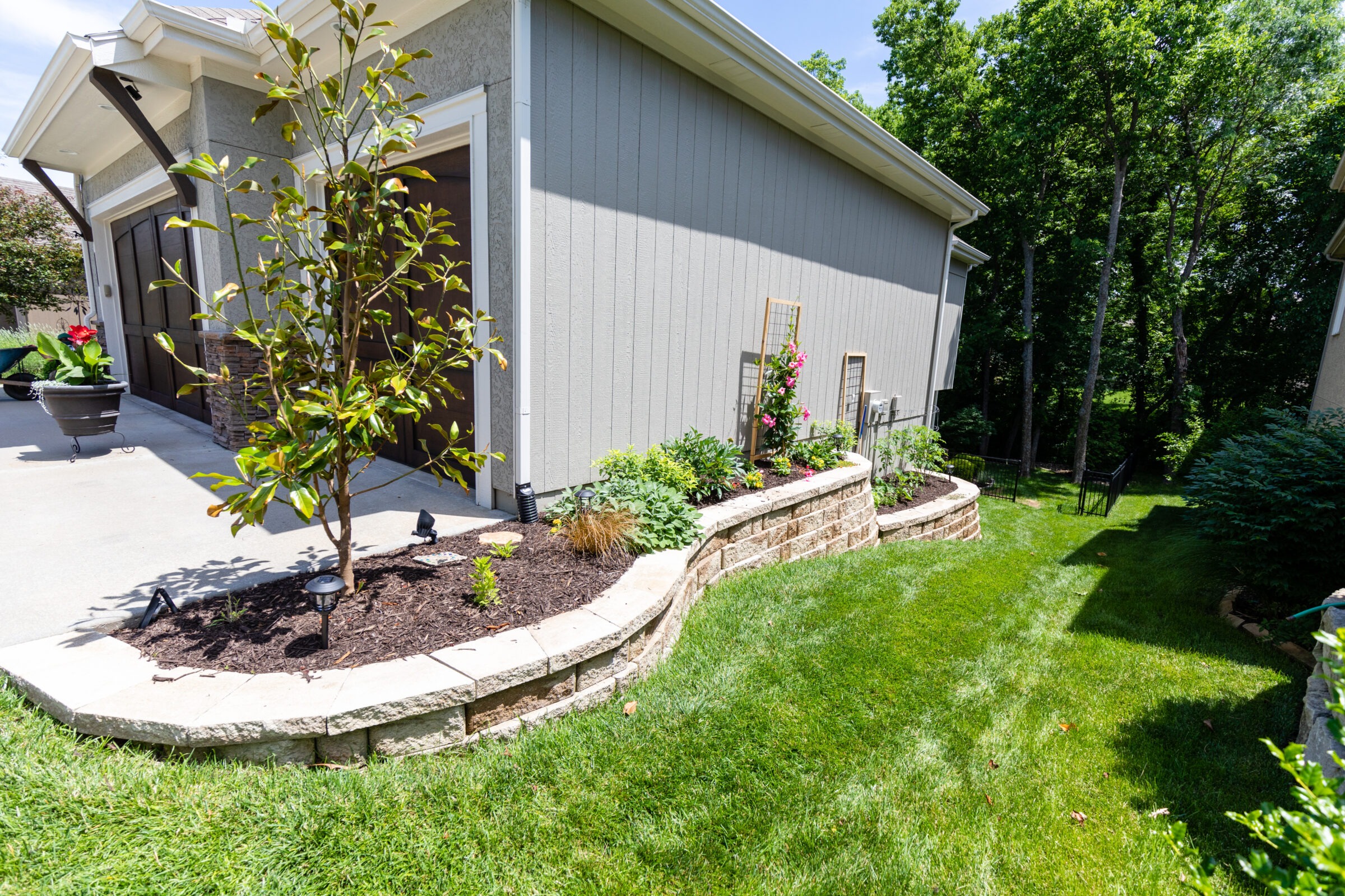 A well-maintained garden beside a house features flowering plants, a small tree, and neatly trimmed grass under clear skies, surrounded by trees.