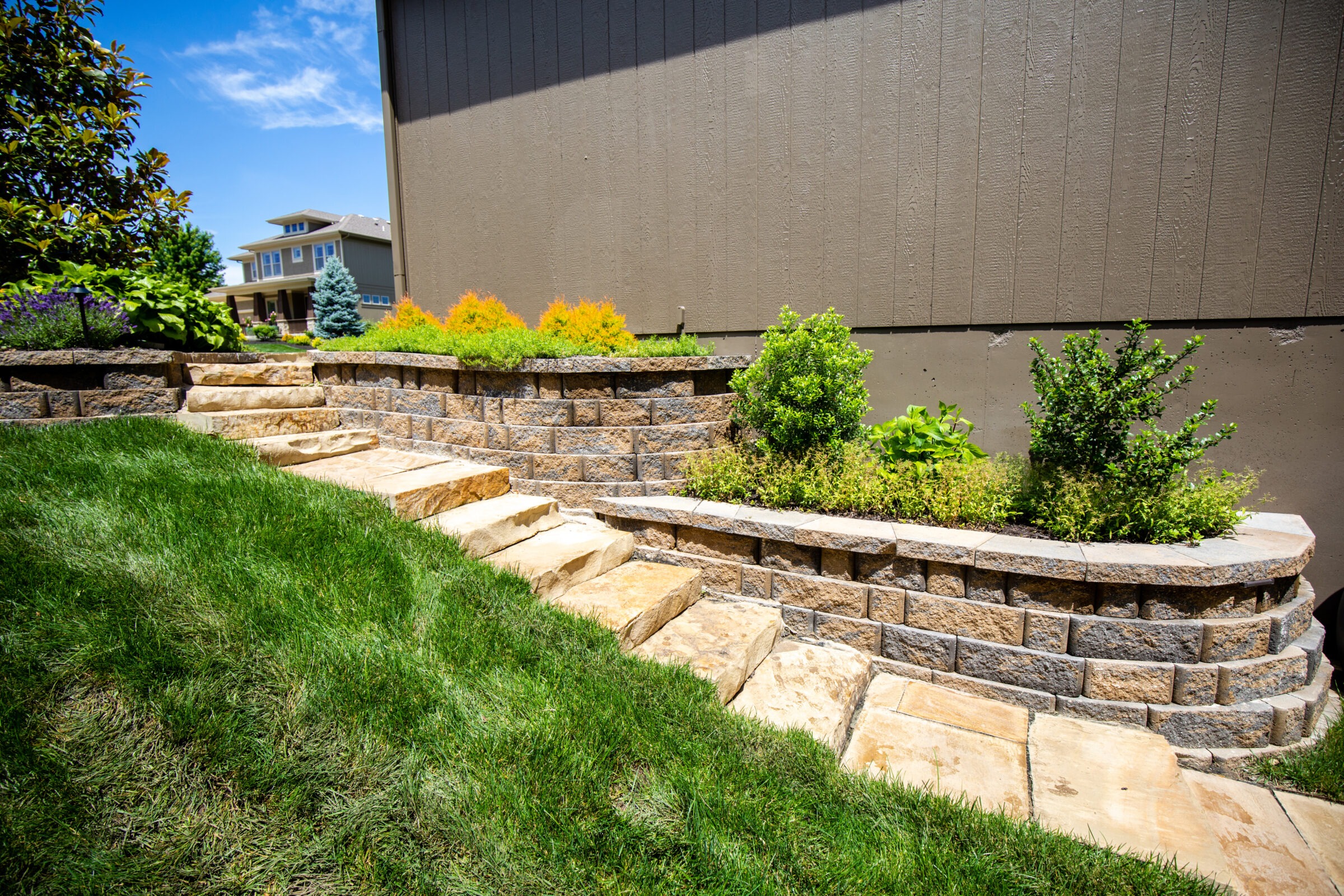 A well-maintained garden with stone steps, retaining wall, and shrubs, leads to a modern house under a clear blue sky.