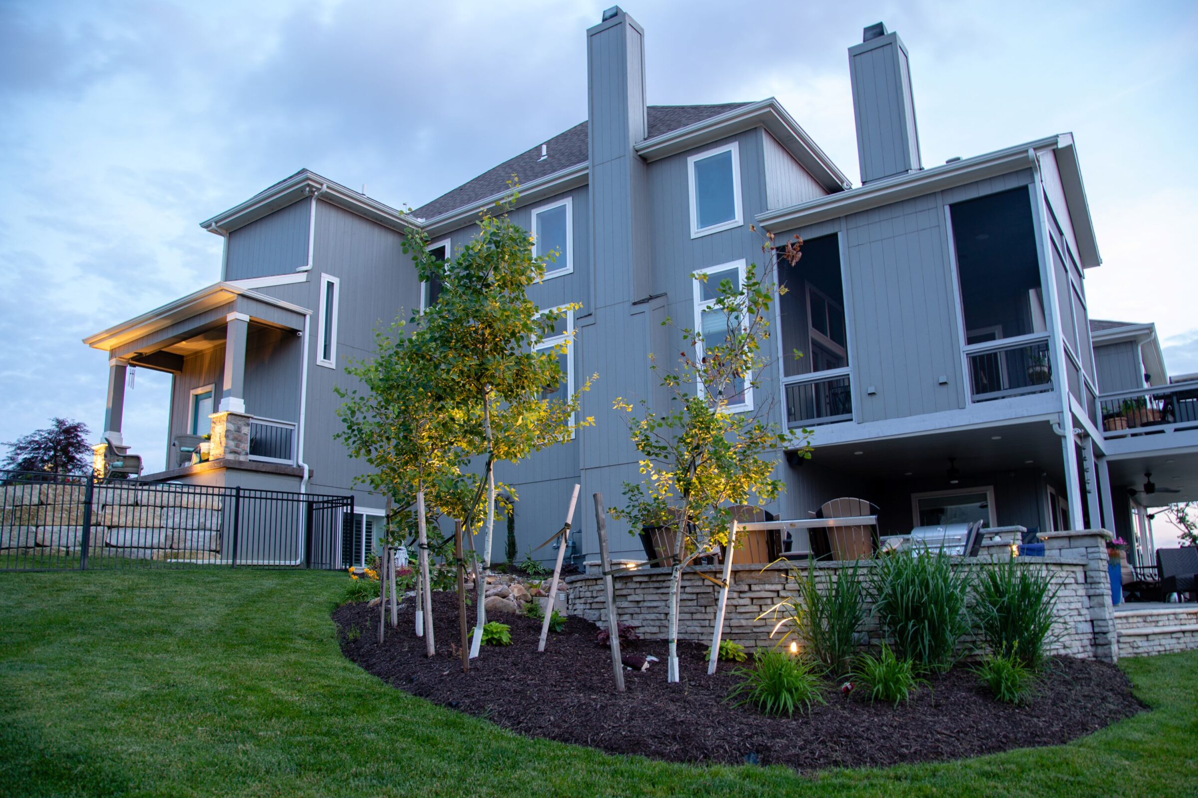 A modern two-story house with large windows and a well-maintained garden, featuring young trees and decorative plants, in a serene setting.