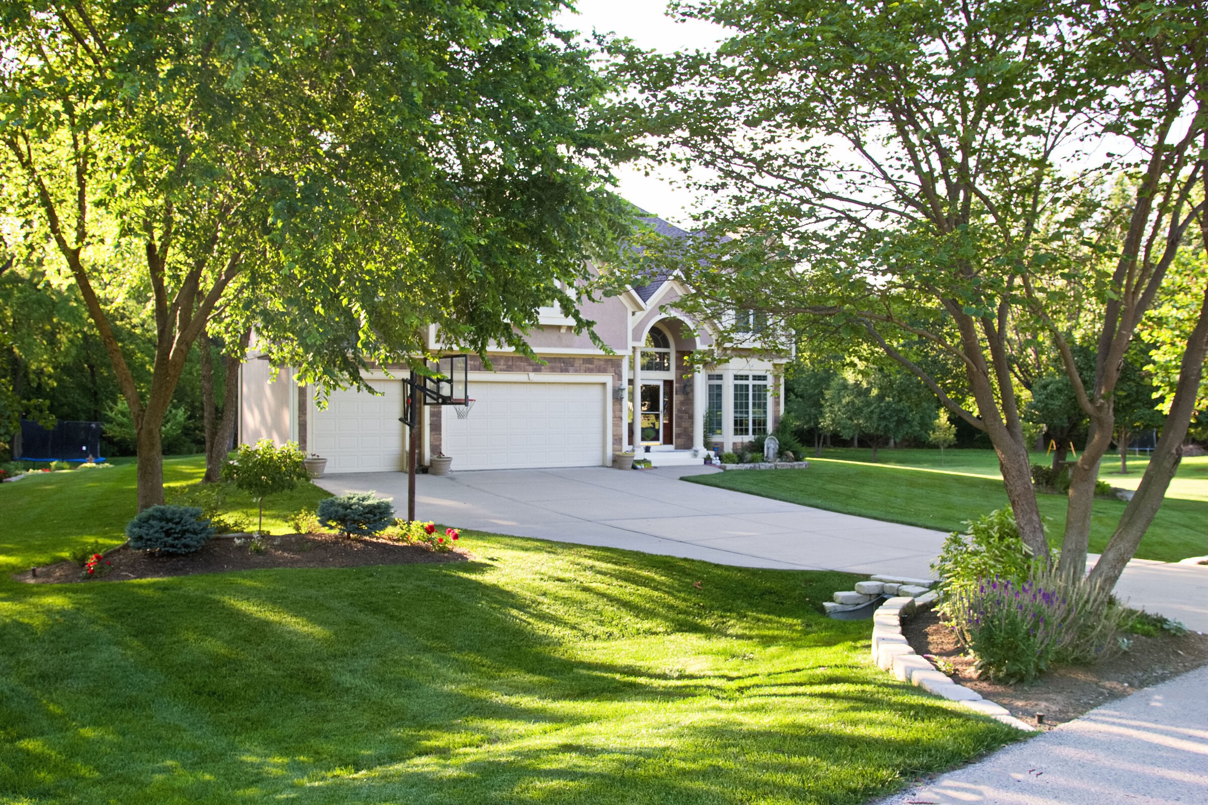 A suburban house with a three-car garage, basketball hoop, and landscaped green lawn surrounded by trees on a sunny day.
