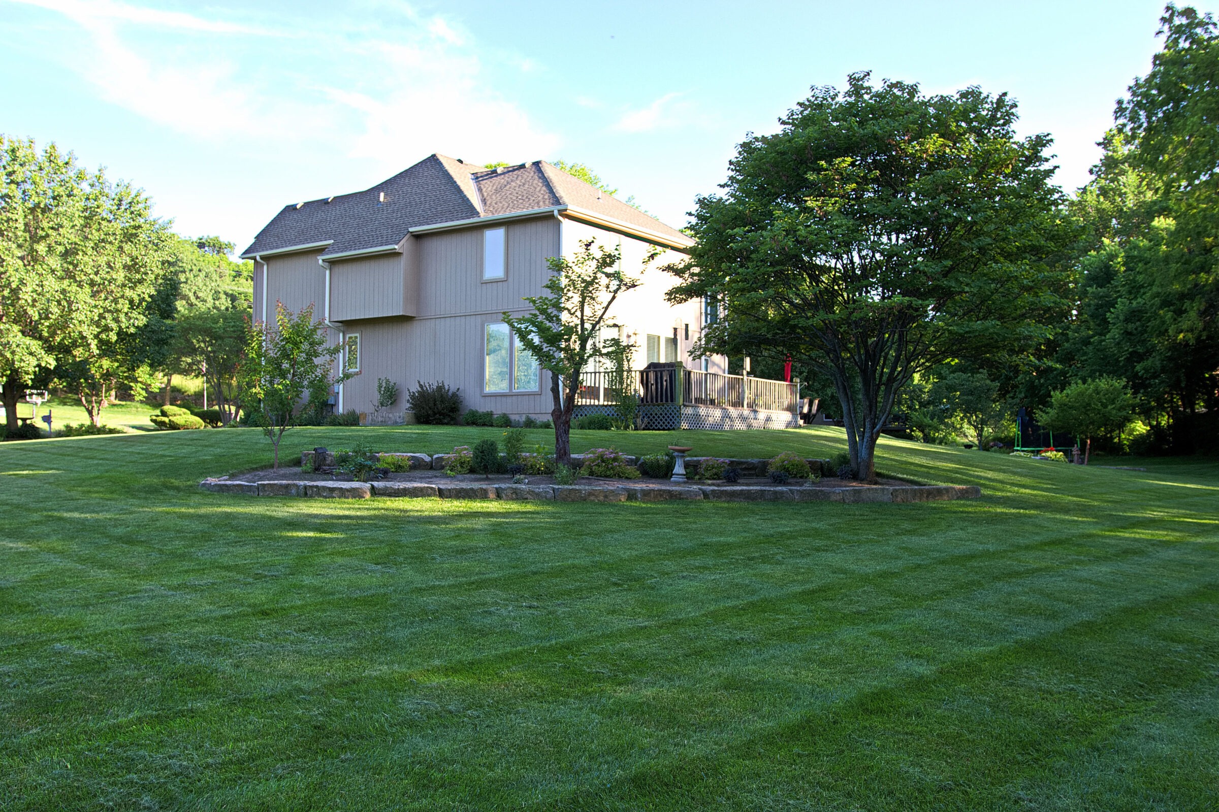 A two-story house surrounded by a well-maintained lawn and trees, with a small garden area in the foreground under clear skies.