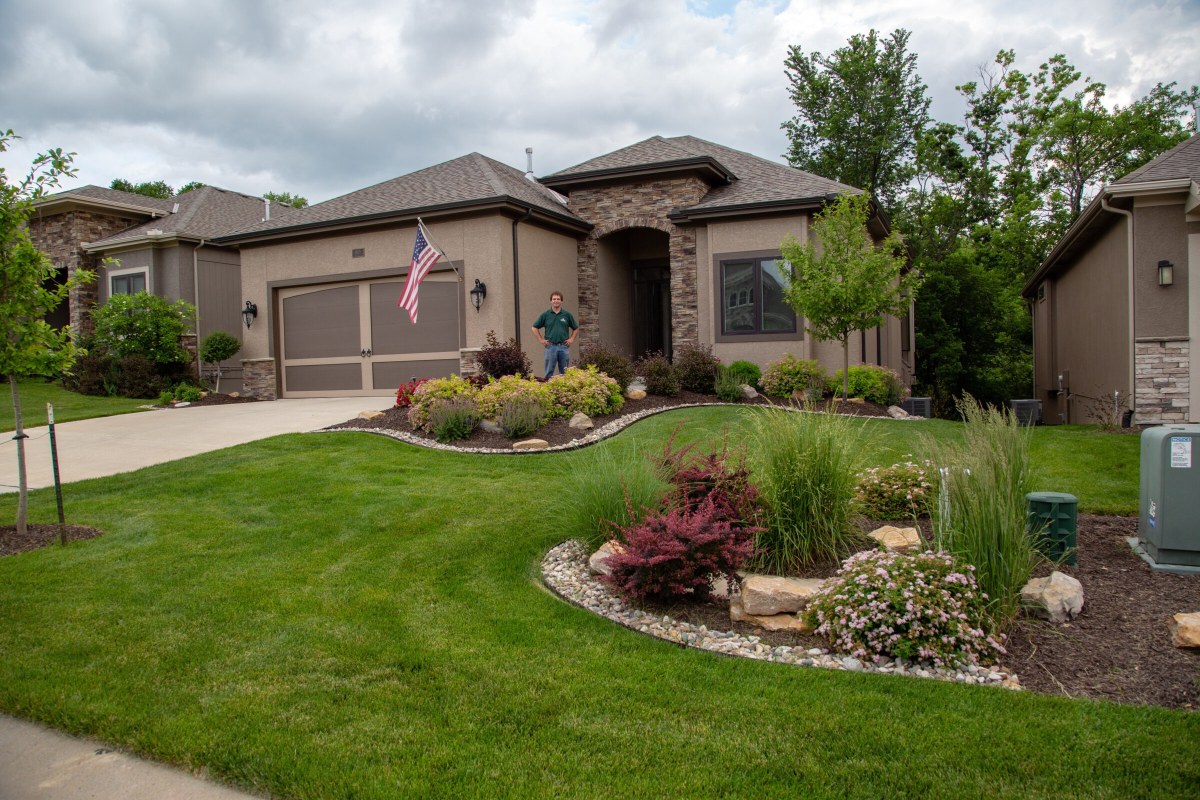 A person stands in front of a suburban house with manicured lawn, garden, and American flag, surrounded by trees and cloudy sky.
