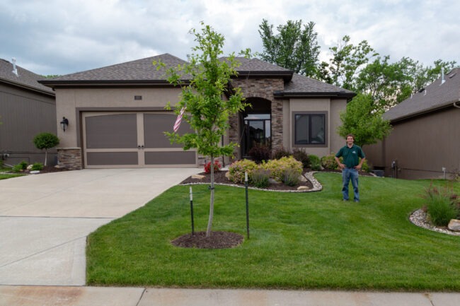 A person stands outside a modern suburban house with a manicured lawn, trees, and stone accents under a cloudy sky.