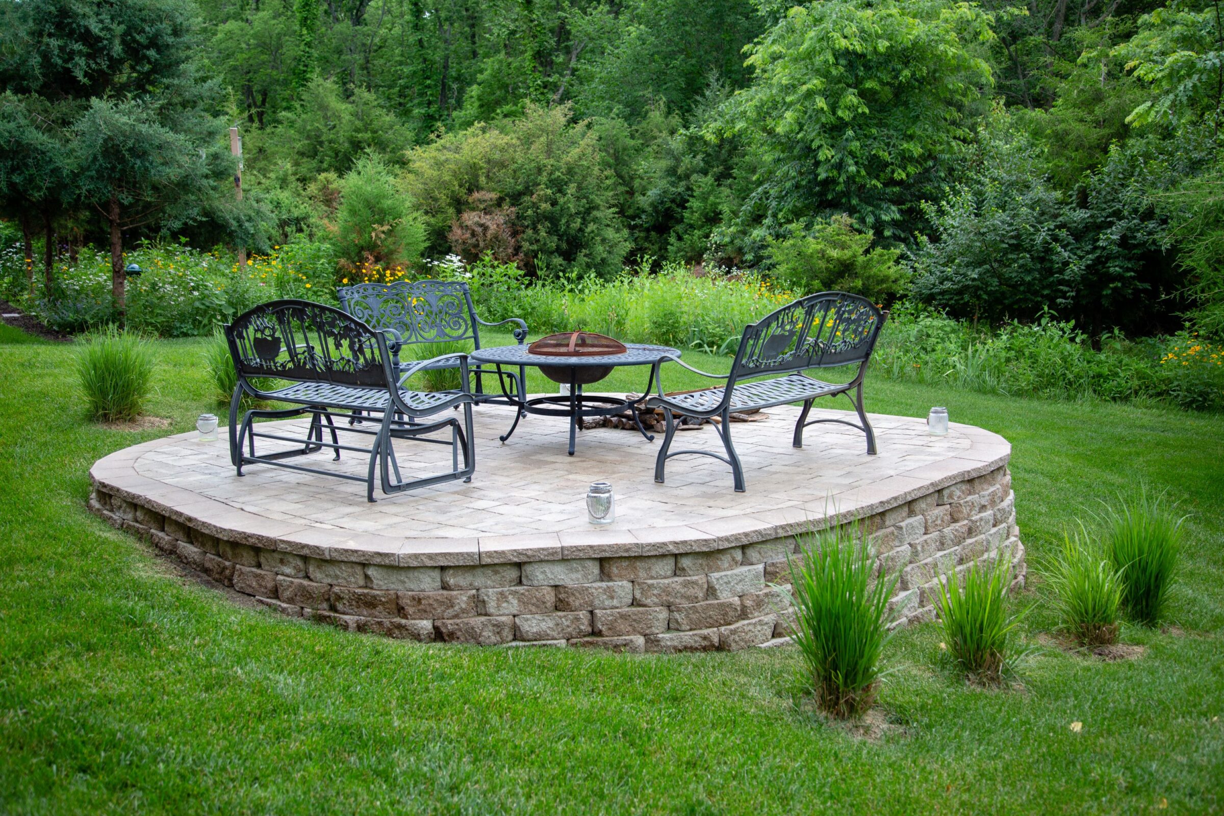 A circular patio with wrought iron furniture, fire pit, surrounded by lush greenery and garden, creating a peaceful outdoor setting.