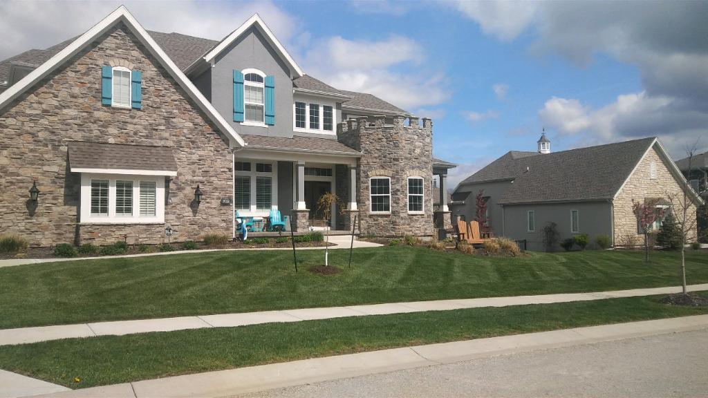 A large, modern stone house with a turret, surrounded by a manicured lawn, under a partly cloudy sky. Cozy neighborhood setting.