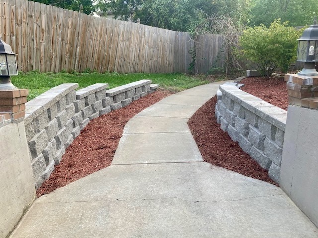 A paved walkway with stone walls, bordered by mulch and grass, leads to a wooden fence, surrounded by lush green foliage.