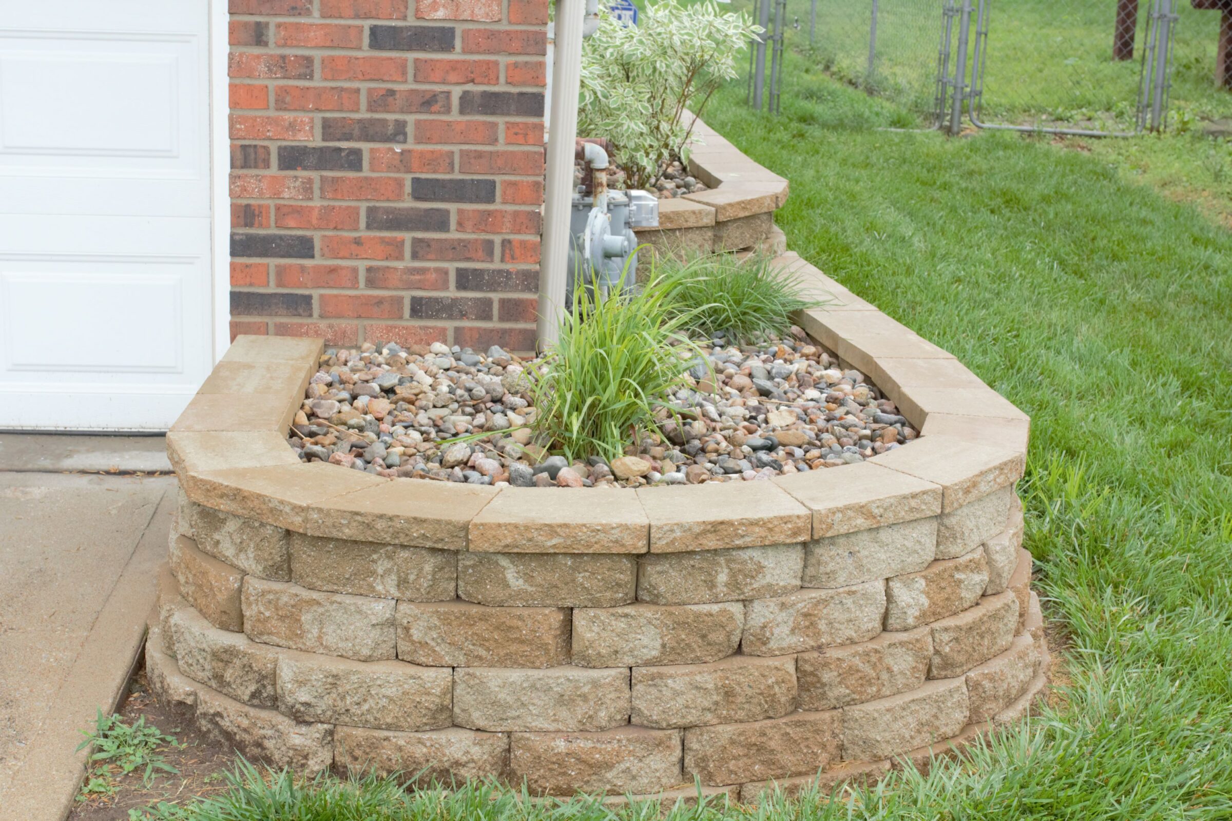 A brick house corner features a small raised garden bed with rocks, plants, and shrubs, bordered by a masonry wall. Grass surrounds it.