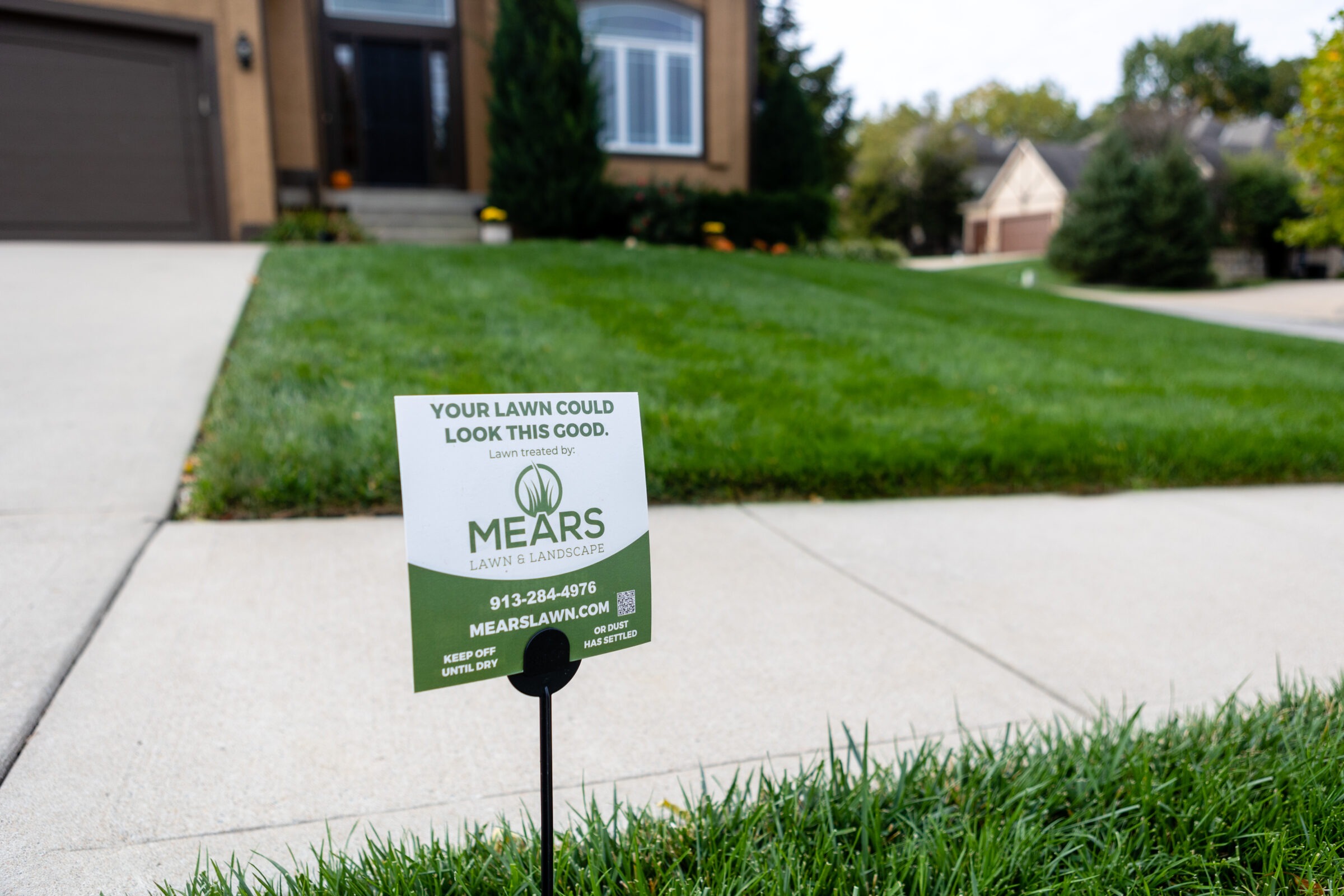 A well-maintained lawn with a sign advertising "Mears Lawn & Landscape," located in a suburban neighborhood with tidy sidewalks and greenery.