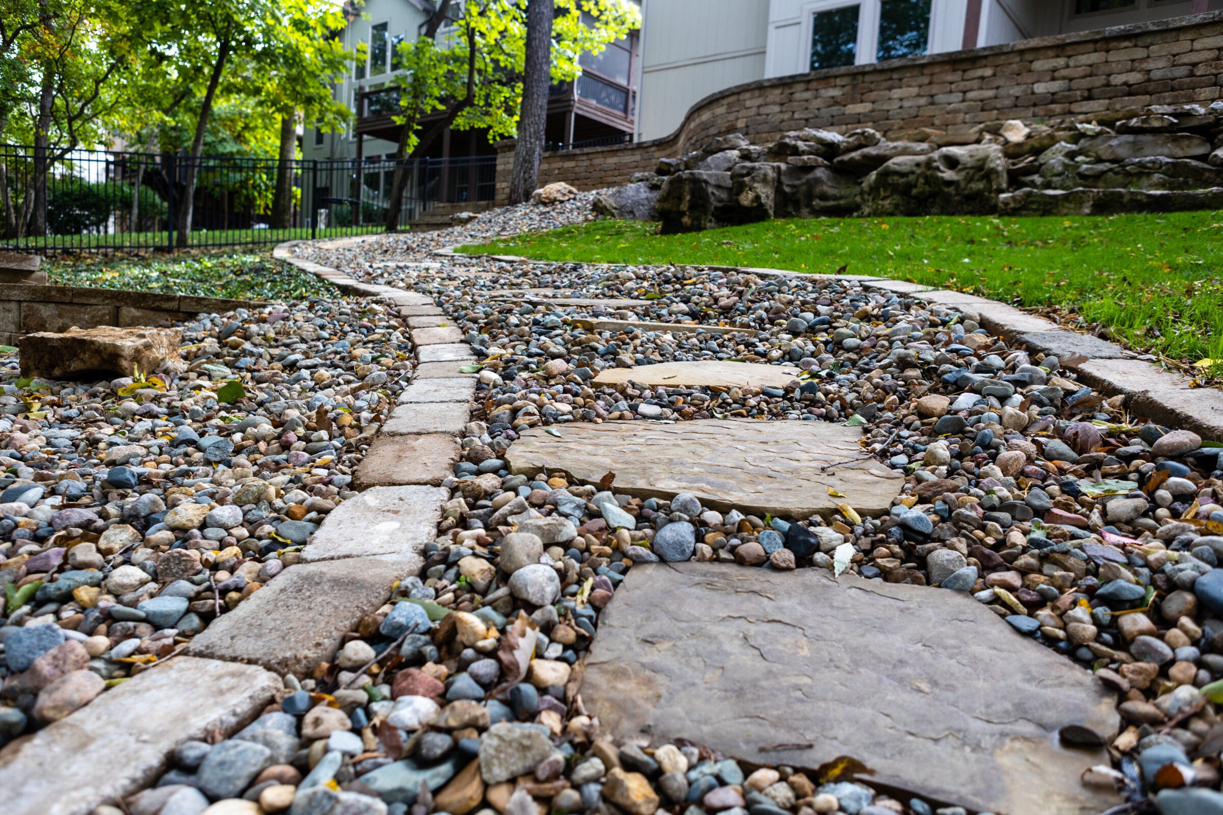 A stepping stone path winds through a garden with trees, surrounded by rocks and greenery, leading towards residential buildings.