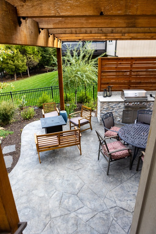 A patio with wooden furniture, a grill, and a stone floor. Surrounded by greenery, wooden pergola, and privacy fence.