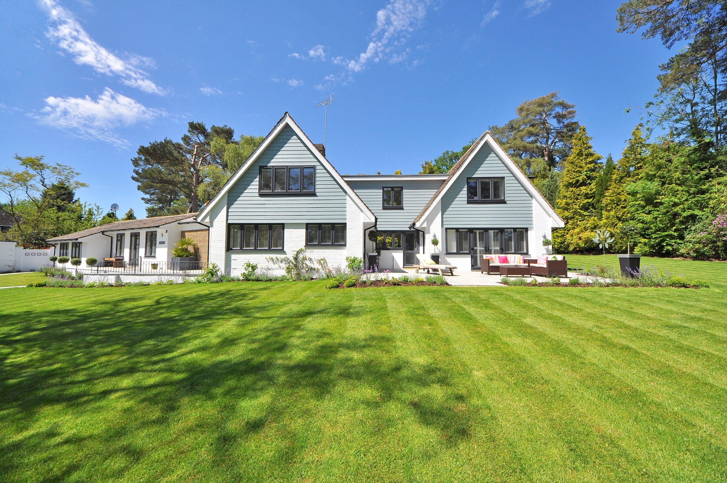A modern house with a large, well-maintained lawn under a clear blue sky, surrounded by tall trees providing shade and greenery.