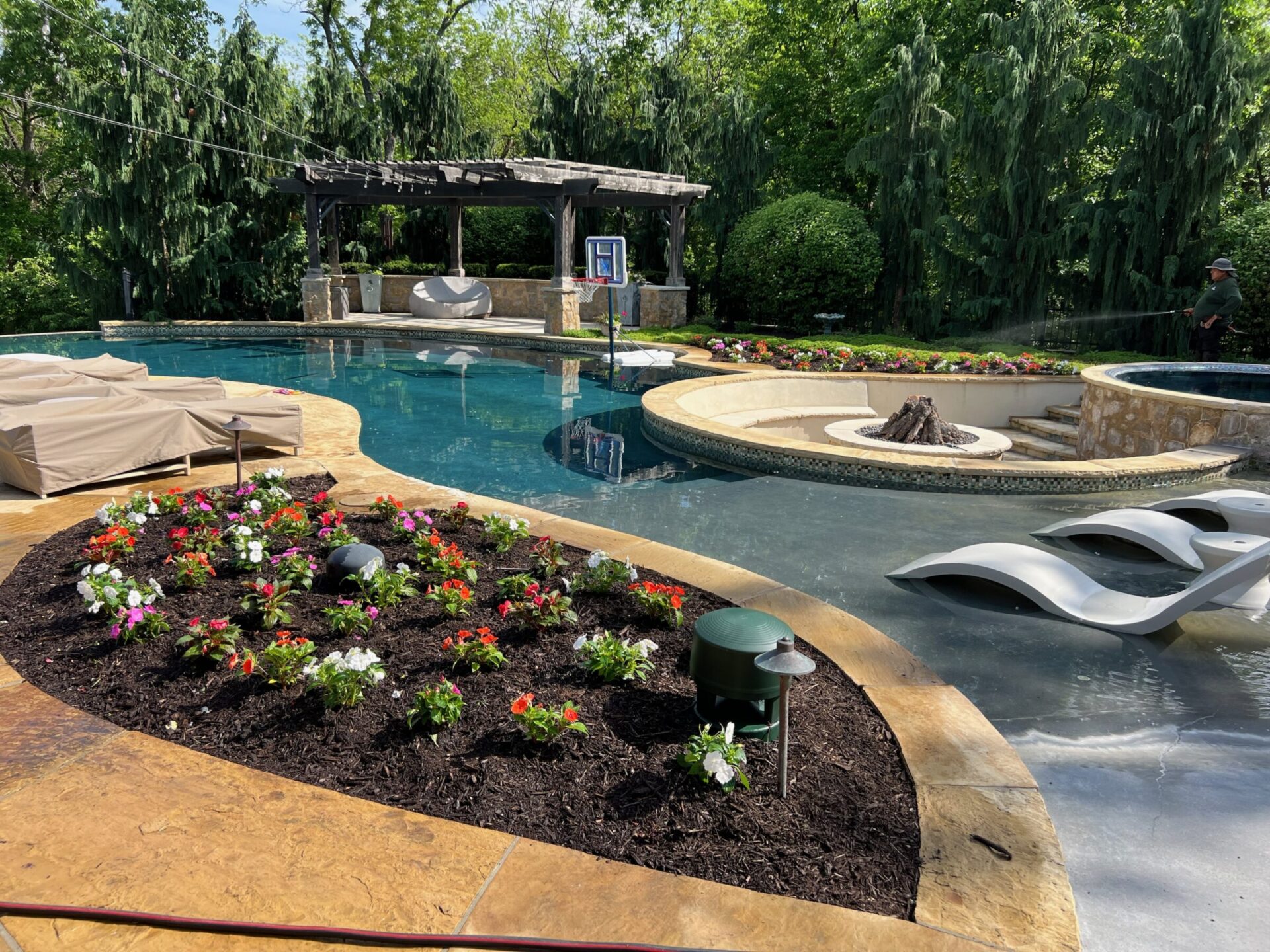 A luxurious backyard pool with a fire pit, pergola, and colorful flower bed surrounded by trees. One person tending the garden.