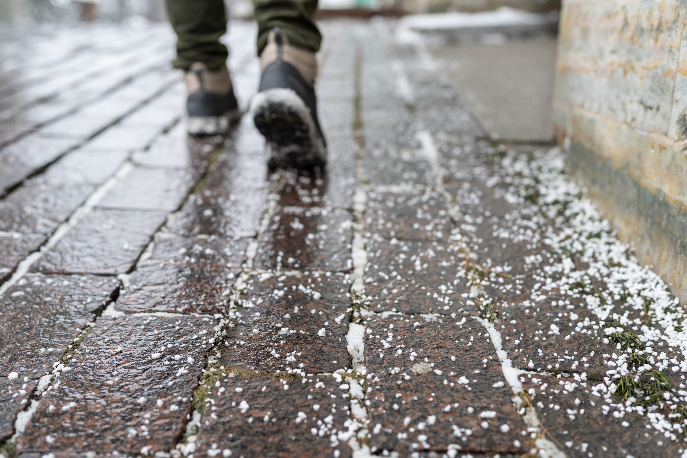A person walks on a wet, brick-paved pathway with small snowflakes scattered, adjacent to a stone wall in a cold environment.