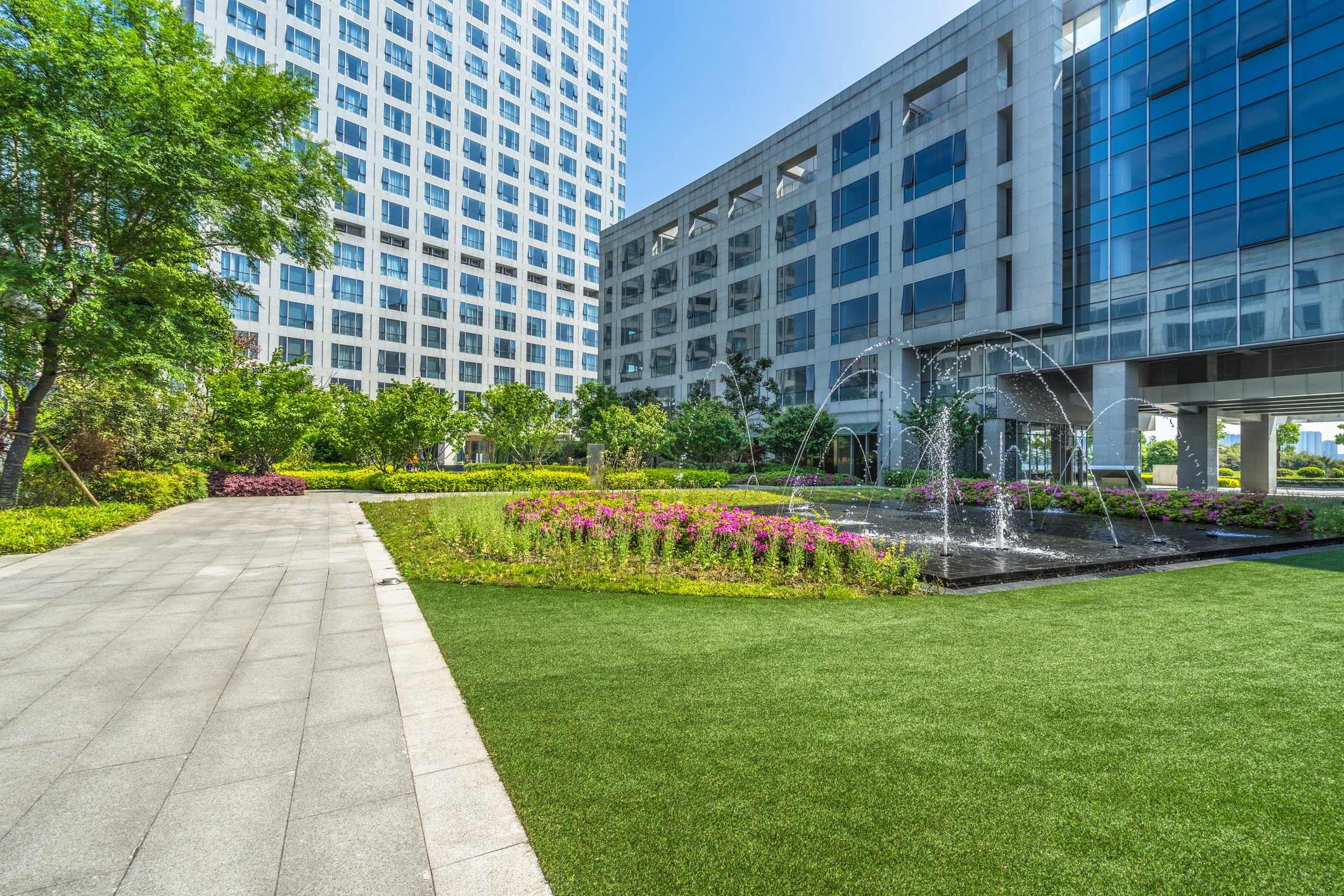 Modern courtyard features lush greenery, colorful flowers, and a fountain, surrounded by tall office buildings under a clear blue sky.
